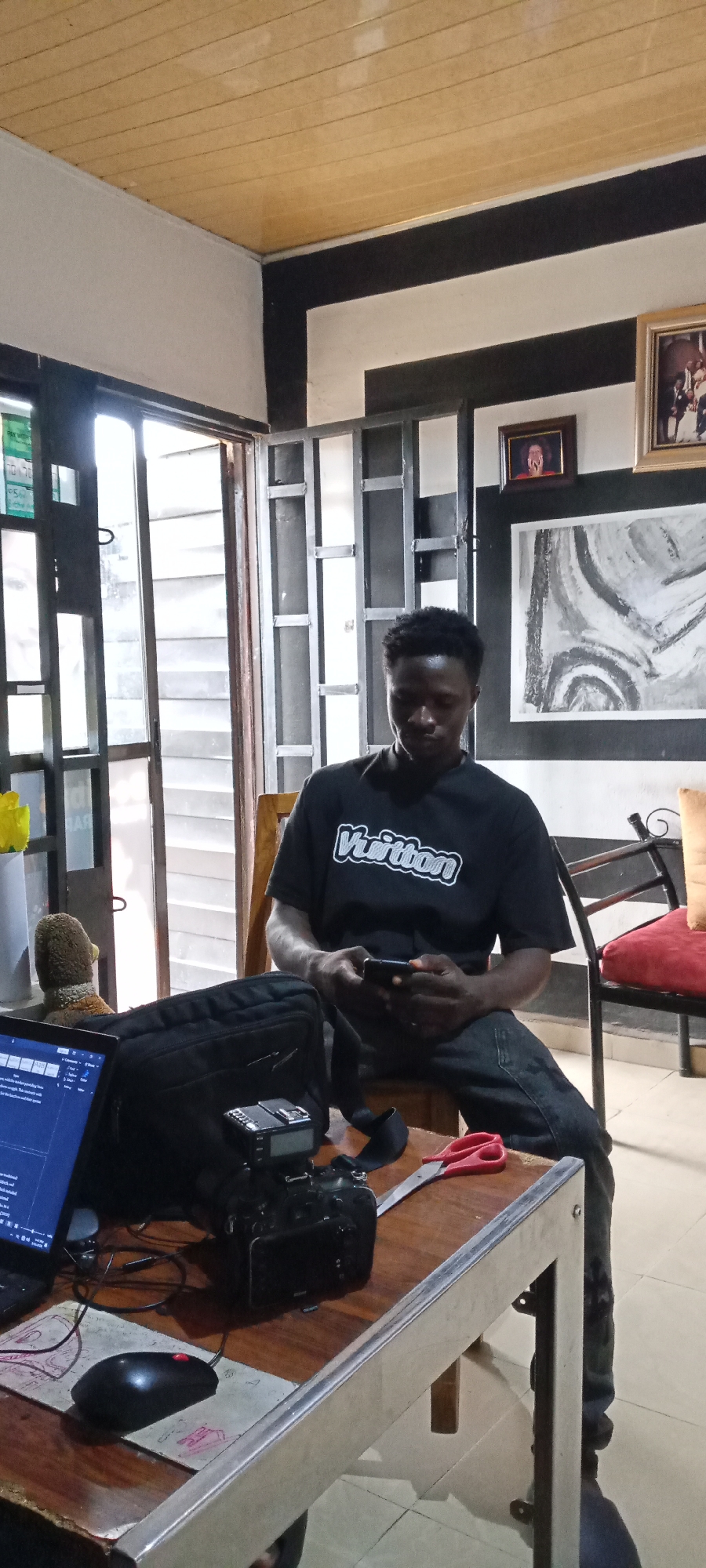 The image shows a person of African descent sitting on a wooden chair in an interior space. The person is wearing a black t-shirt with the word "Vuitton" printed on it and dark jeans. They are looking down at a mobile phone in their hands.
On a table in front of the person are a laptop, a camera with a flash unit, a black bag, a mouse on a mousepad, and a pair of red-handled scissors. The room has black and white striped walls and a wooden ceiling. A window with a security gate is visible to the left. There are framed pictures on the wall. There is a red cushion placed on a chair next to the person.
The location is identified as the city of Unknown, Unknown.