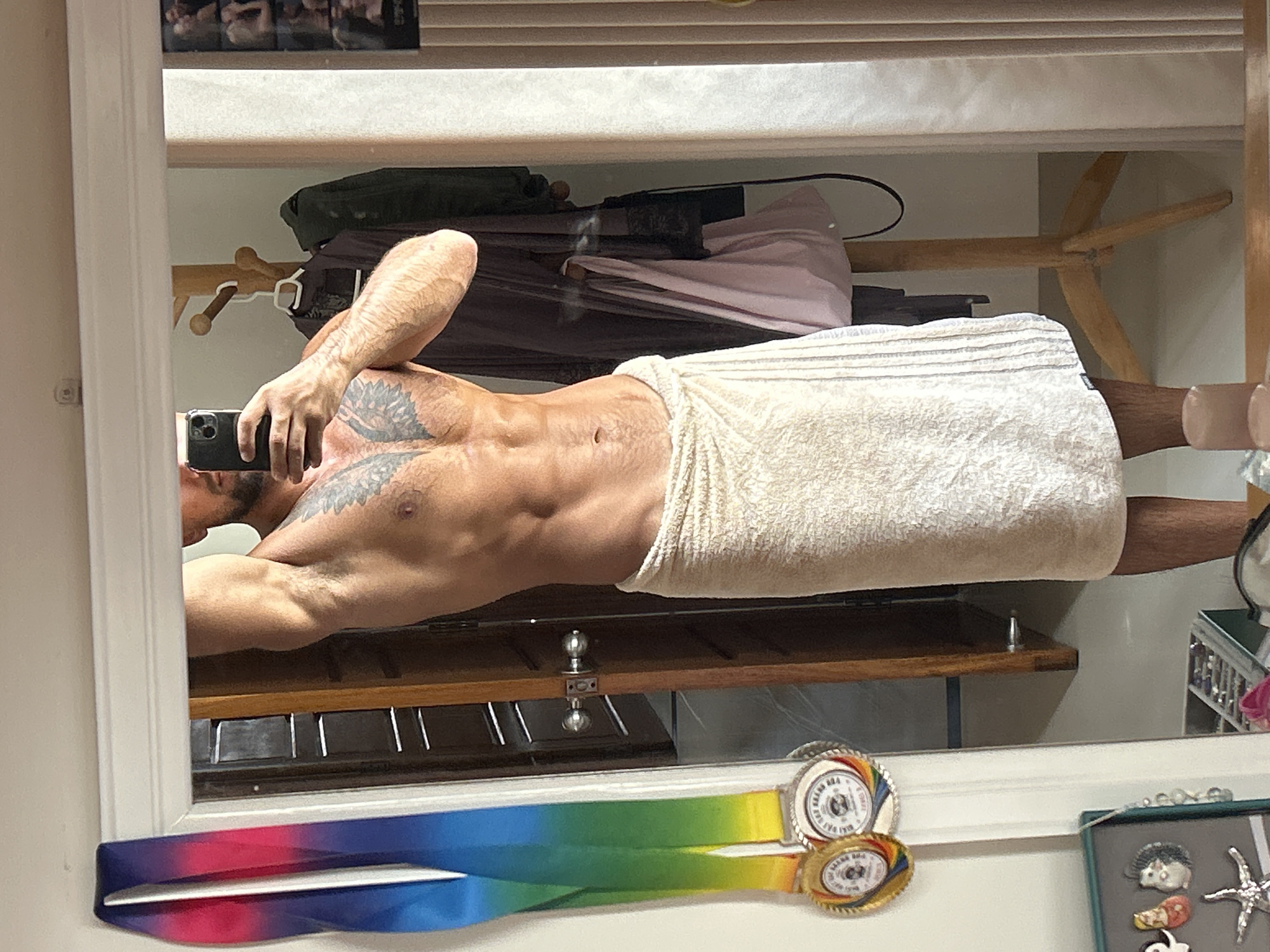In a bathroom in Nha Trang Ward, Vietnam, a man is taking a mirror selfie. He is holding a smartphone in his right hand and is wearing a towel wrapped around his waist. He has a tattoo on his chest and visible abdominal muscles. A rack with clothes hangs in the background. On the left side of the mirror, two medals are displayed on ribbons. In the lower left corner, there are decorative items and a jewelry box.