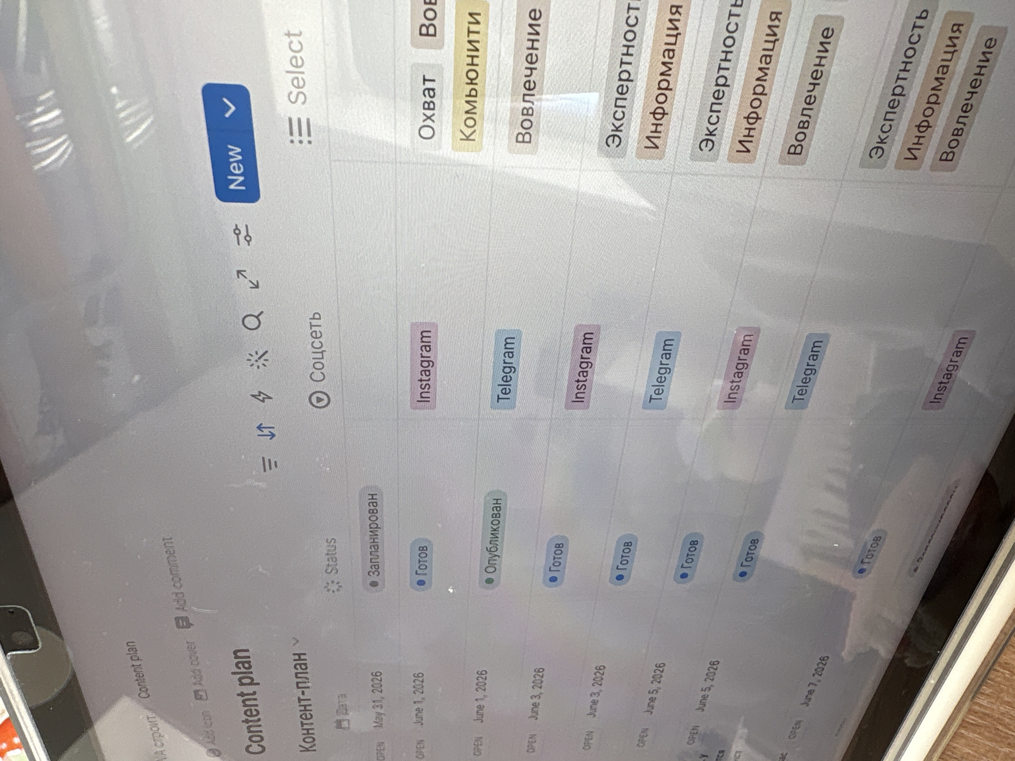 The image shows a close-up of a tablet screen displaying a content plan table. The table includes columns for date, status, social network (Instagram, Telegram), and metrics like reach, community, and engagement. The content plan is in Russian, with status options including "Planned," "Ready," and "Published." The date column shows dates from May 31, 2026, to June 7, 2026. The tablet is resting on a surface with a wooden texture. The location is not revealed in this image.