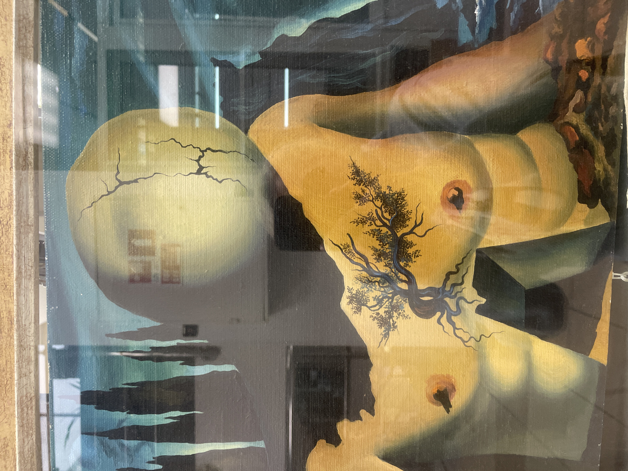 The image is a painting, framed in a light brown wooden frame. The painting depicts a surreal landscape with a disembodied torso in the foreground. The torso is rendered in a yellow-toned flesh color and has a tree growing from its chest. The background features dark, spiky shapes resembling trees or rock formations against a teal sky. To the right, a dark, craggy mountain or rock face descends into dark, churning water. A large, pale, cracked sphere is positioned above and behind the torso, appearing to be the head. The overall style of the painting is reminiscent of Salvador Dalí. The painting is behind glass, as evidenced by reflections of indoor lighting and what appear to be ceiling lights or fixtures. There is no discernible location in a city or any specific environmental details that would indicate its placement.