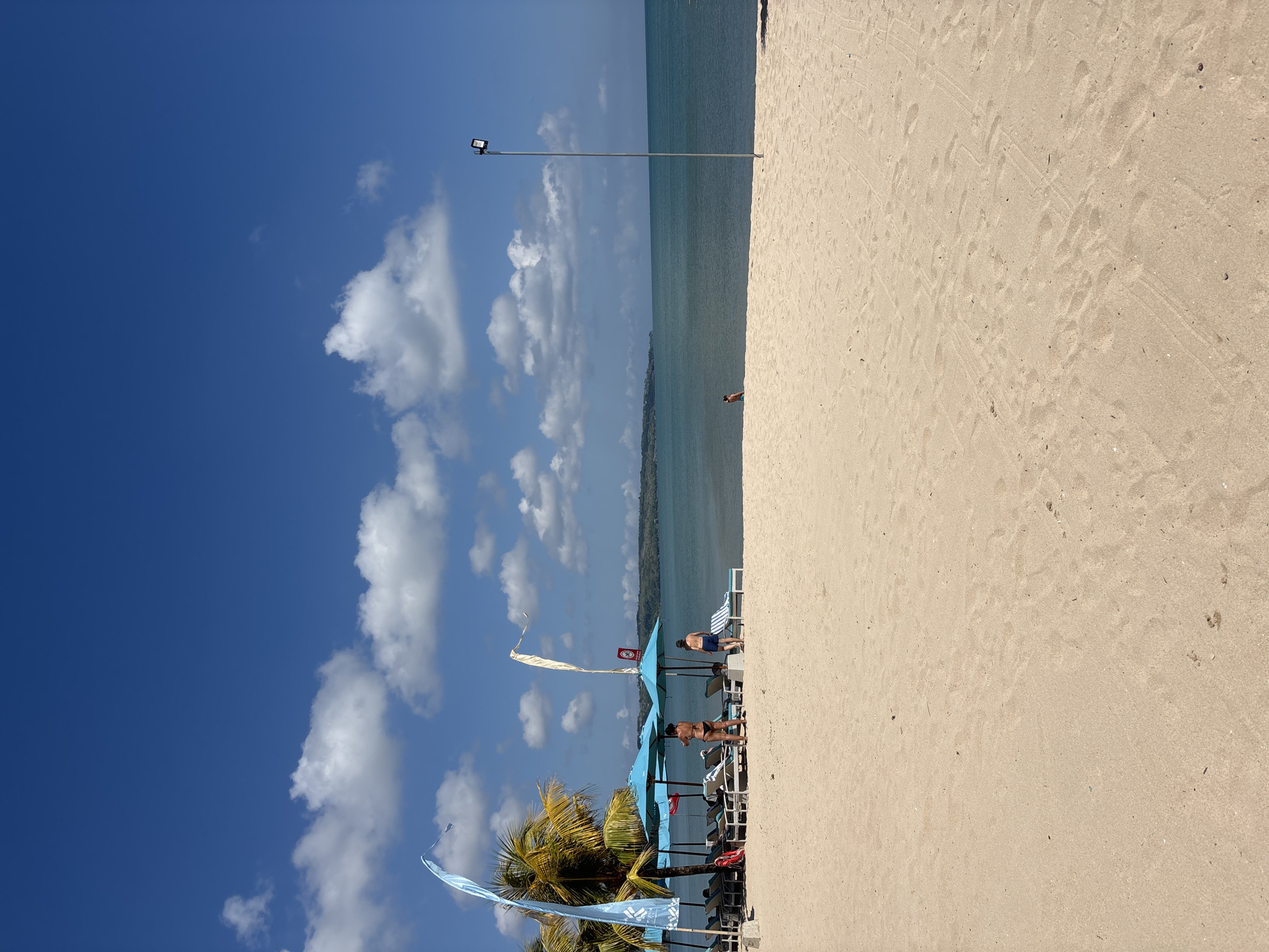 This is a photograph of a sandy beach in Kuta, Indonesia, on a clear day with a bright blue sky dotted with white clouds. In the foreground, the sand is smooth with some footprints. In the middle ground, a line of lounge chairs and blue umbrellas is set up near palm trees. Two individuals are standing near the umbrellas, and a third person is walking further down the beach. The ocean is visible in the background, stretching towards a distant landmass. A tall pole with a spotlight stands on the beach to the right.