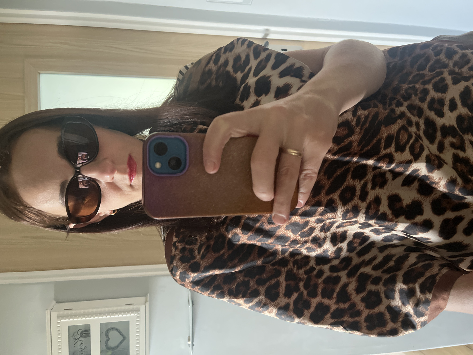 An overhead, mirrored selfie captures a person holding a smartphone, facing the camera, in what appears to be an indoor setting. The image is oriented with the person's head to the left, but is effectively a portrait selfie taken in a mirror.
The main subject is a person, likely female, with long dark hair, wearing large dark sunglasses and red lipstick. A distinct red lipstick kiss mark is visible on her left cheek/jawline. She is wearing a garment with a prominent leopard print pattern. On her right hand, holding the phone, a simple gold band ring is visible on her ring finger.
The smartphone she is holding has a purple-blue camera module and what appears to be a shimmering or textured brown/gold case. The phone is angled, with her fingers gripping the side and back.
The background, visible as a reflection in the mirror, shows part of a room. Features include light-colored walls, a wooden doorframe, and a white decorative frame or picture frame mounted on a lower wall section. Within this frame, text that appears to read "Love" and a heart symbol can be faintly seen. The lighting suggests it is daytime.