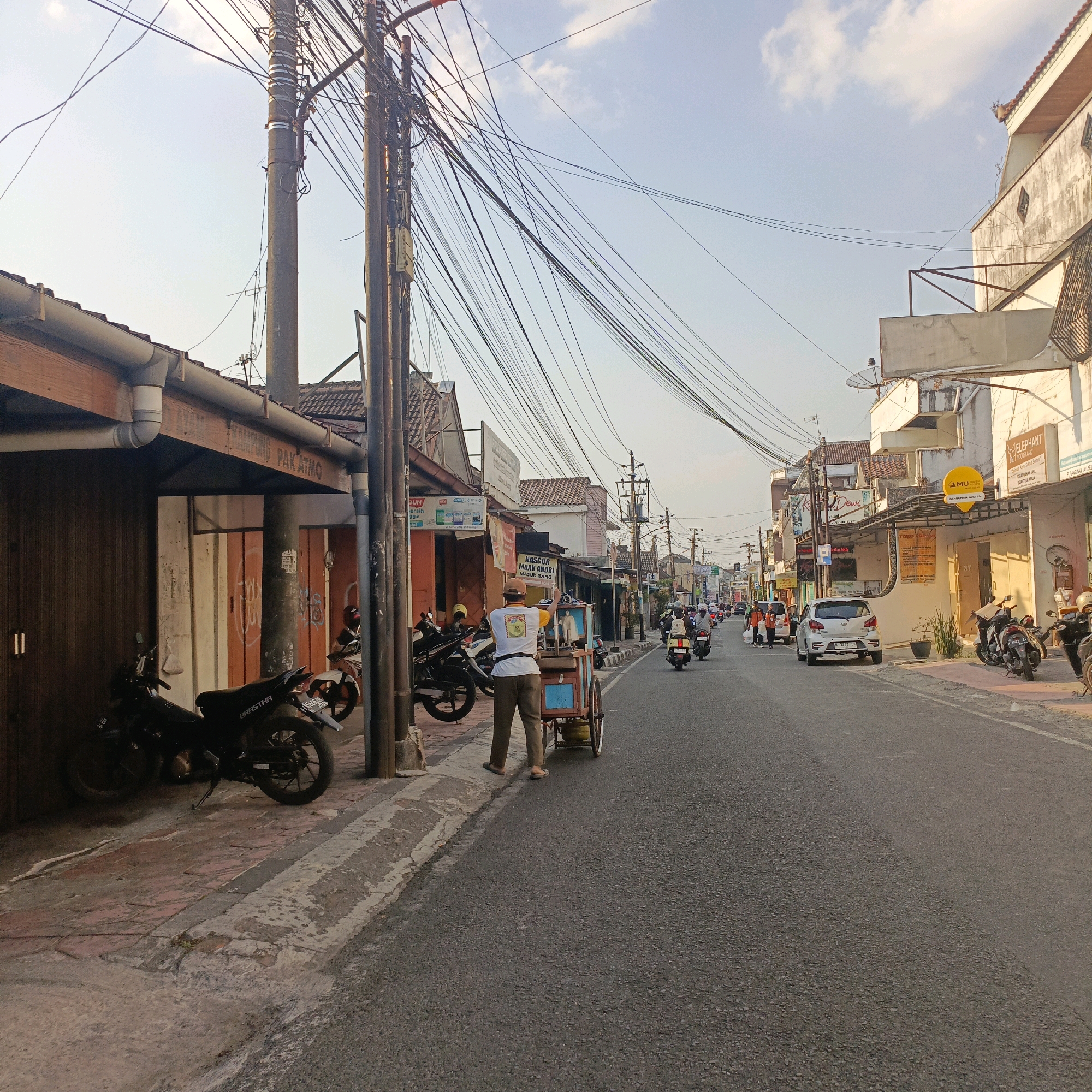 The image depicts a street scene in Salatiga, Indonesia, likely in the afternoon due to the lighting. The street is paved with asphalt and lined with buildings on both sides. Overhead, a complex network of electrical wires and poles dominates the sky.
On the left side of the street, a row of buildings features brown wooden doors and some signage, including one that reads "PAK ATMO". Several motorcycles are parked along the sidewalk in front of these buildings. A person is standing near a small cart, holding a sign that is partially visible and reads "NASGOR MBAK ANDRI MASUK GANG".
On the right side of the street, there are more buildings, some appearing to be shops or businesses. One building has a sign for "ELEPHANT WOODPLANK". Another sign is a yellow circular "MU" logo. Several more motorcycles are parked along the sidewalk. A silver car is visible in the middle of the street, and other motorcycles are moving along the road. The overall impression is of a busy urban environment with a mix of commercial activity and everyday life.