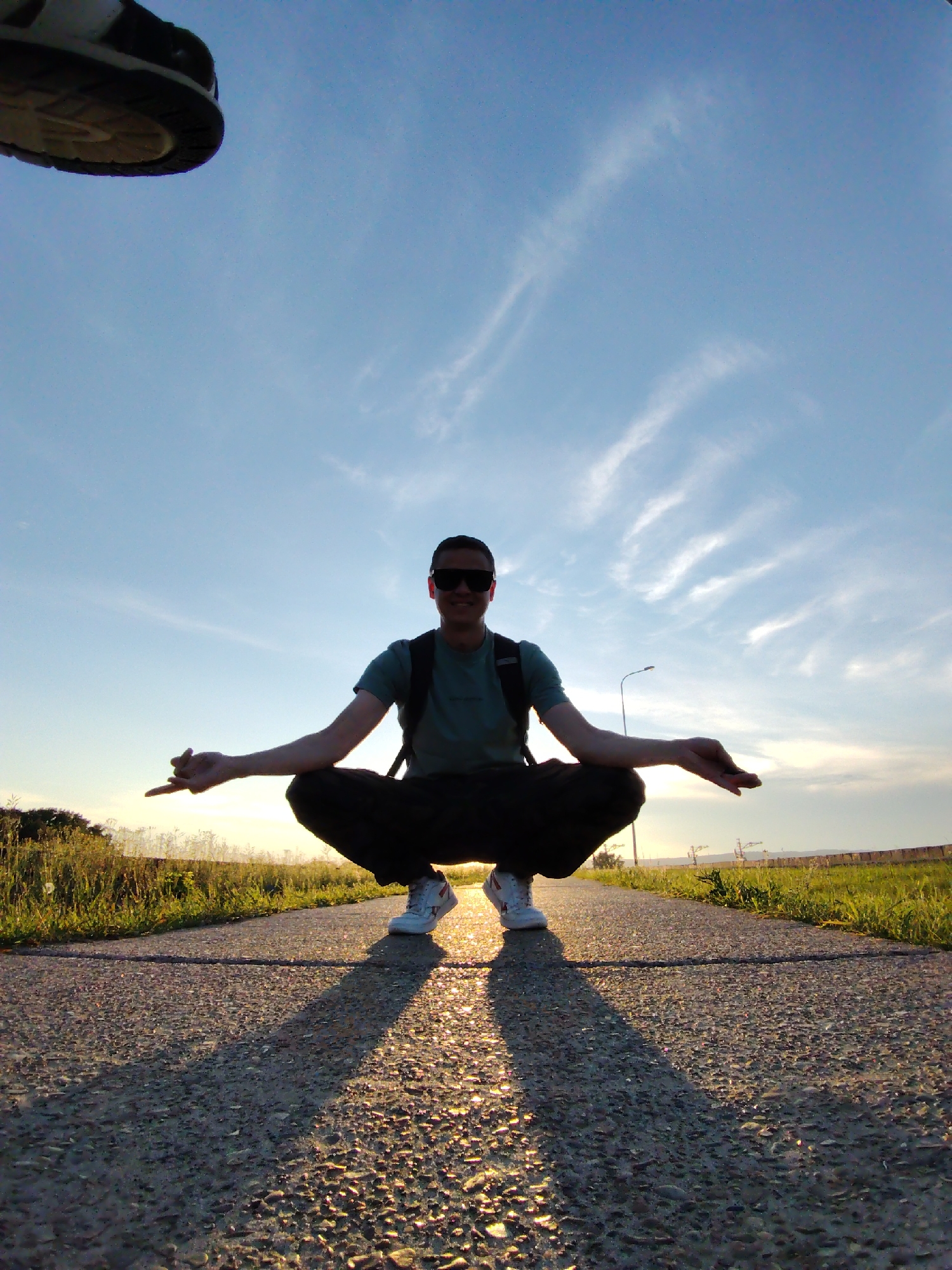 An adult male is captured from a low angle, squatting on a paved path during a sunset. The man is wearing sunglasses, a dark t-shirt, and dark pants, with a backpack on his shoulders. His arms are extended outwards, with his left hand in a gesture resembling a pinch between his thumb and index finger, and his right hand open, palm up. The path appears to be a road or sidewalk, bordered by dry grass and vegetation on either side. The sky is a clear blue with wispy white clouds, illuminated by the warm light of the setting sun, casting long shadows on the path. In the background, a lamppost and some distant structures are visible. The composition suggests the image was taken in an open, outdoor area, potentially on the outskirts of a populated area. The presence of a lamppost and distant structures could indicate proximity to a town or city. Without further context, it is not possible to definitively place this scene in Chaikovsky, Russia, as similar environments can be found in many locations.