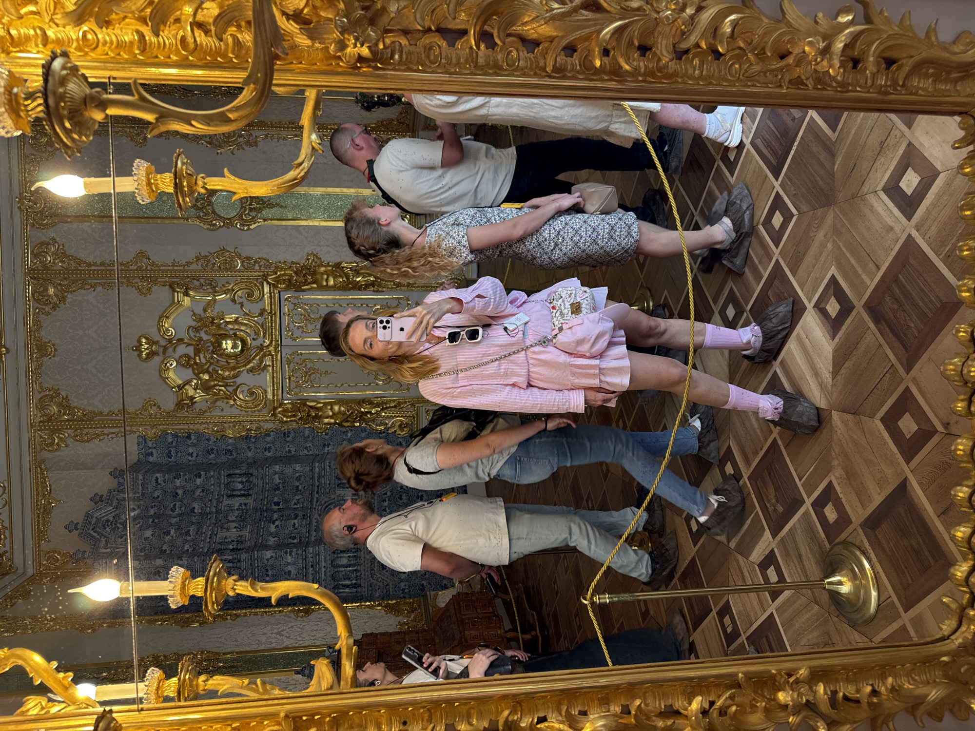 In the Catherine Palace in Pushkin, Russia, a woman in a pink dress and fuzzy pink socks takes a selfie in an ornate gold mirror. She is positioned in the foreground, with her phone held up to capture her reflection. Behind her, several other people, some wearing protective shoe covers, walk through the hall. The hall features ornate gold decorations on the walls and ceiling, a patterned wooden floor, and a tiled blue wall. The mirror itself is elaborately framed in gold.