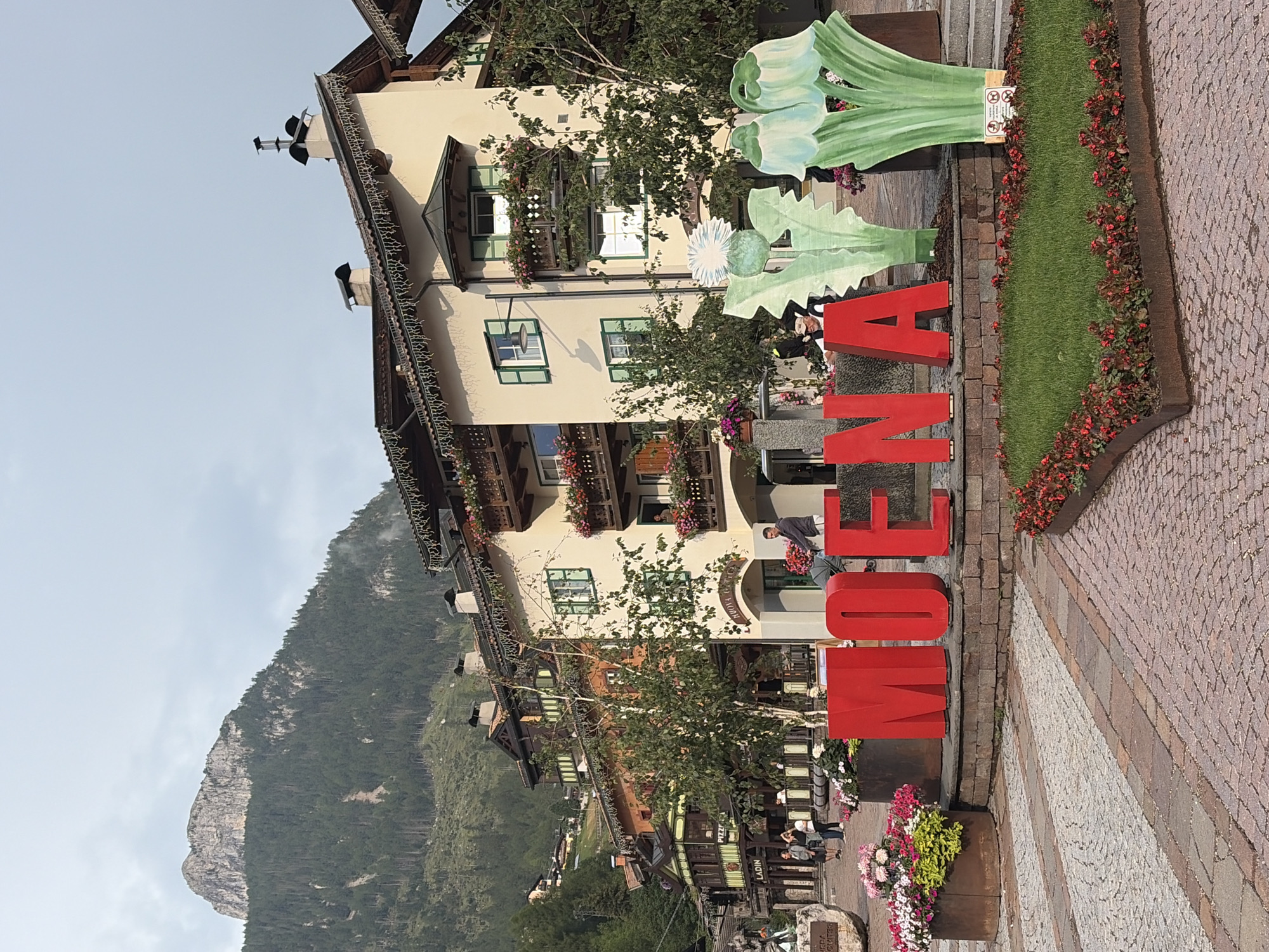 The image depicts a street scene in Moena, Italy, featuring large red letters spelling out "MOENA" in the foreground. Behind the letters, there are a series of multi-story buildings with traditional alpine architecture, characterized by wooden balconies adorned with flower boxes and decorative lights along the eaves. A steep, tree-covered mountain rises in the background under a cloudy sky. In the middle ground, a few people are visible, including a man pushing a stroller and others walking. A grassy area with red flowers borders the "MOENA" letters. The street itself is paved with cobblestones. The overall scene suggests a charming town square in a mountainous region, likely during daytime, with a focus on local identity through the prominent display of the town's name.