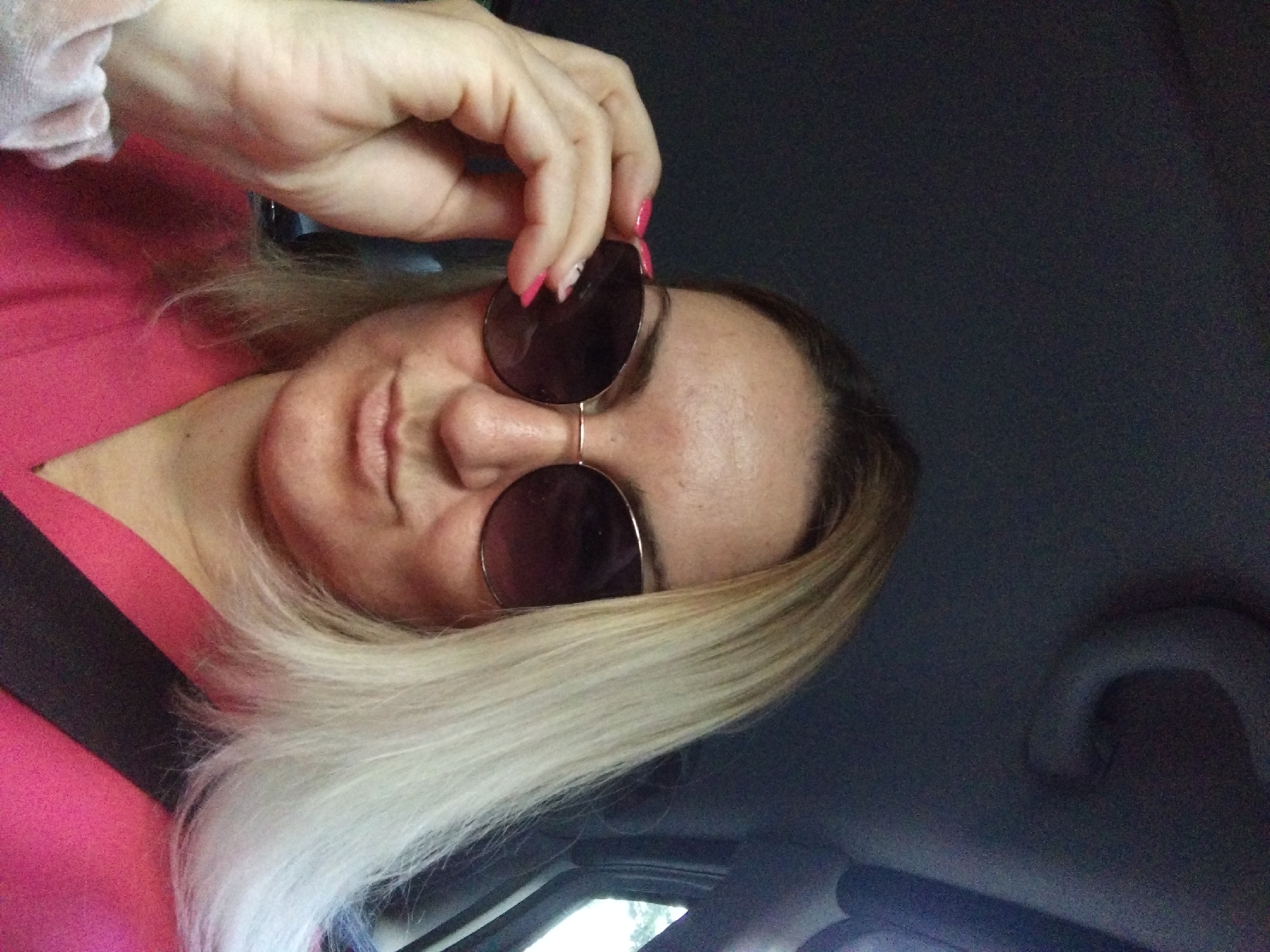 A woman is in a car, possibly driving or a passenger. She is wearing sunglasses and a pink top. She is holding her sunglasses with her left hand. The interior of the car, including the roof and seatbelt, are visible. The location is unknown.