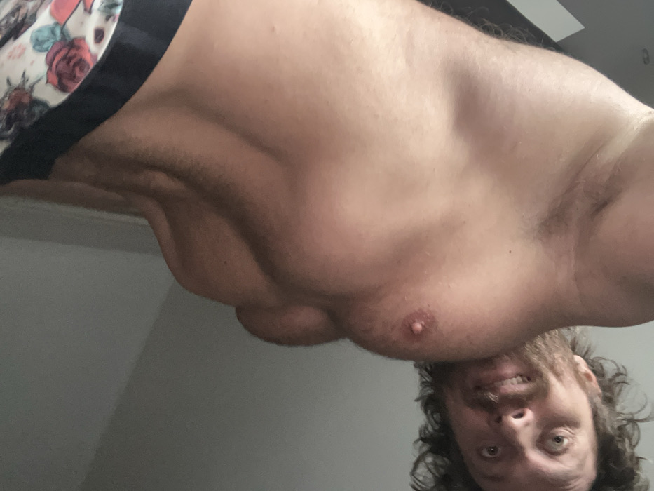 A male individual with light skin, curly brown hair, and facial hair is depicted from a low-angle perspective. He is shirtless, revealing a muscular physique across his chest and abdomen, with the right pectoral muscle prominently visible. His left armpit and side are also visible. He wears patterned boxer briefs featuring a floral design of red roses on a light background, with a dark waistband. His head is tilted, and he looks directly into the camera with wide eyes and a slight grimace or grin that exposes his upper teeth. The background consists of a plain, light grey wall or ceiling, with a dark, vertical architectural element present on the far left side of the frame. The scene's specific location within Saint Petersburg, Russia, is not discernible from the provided visual information.