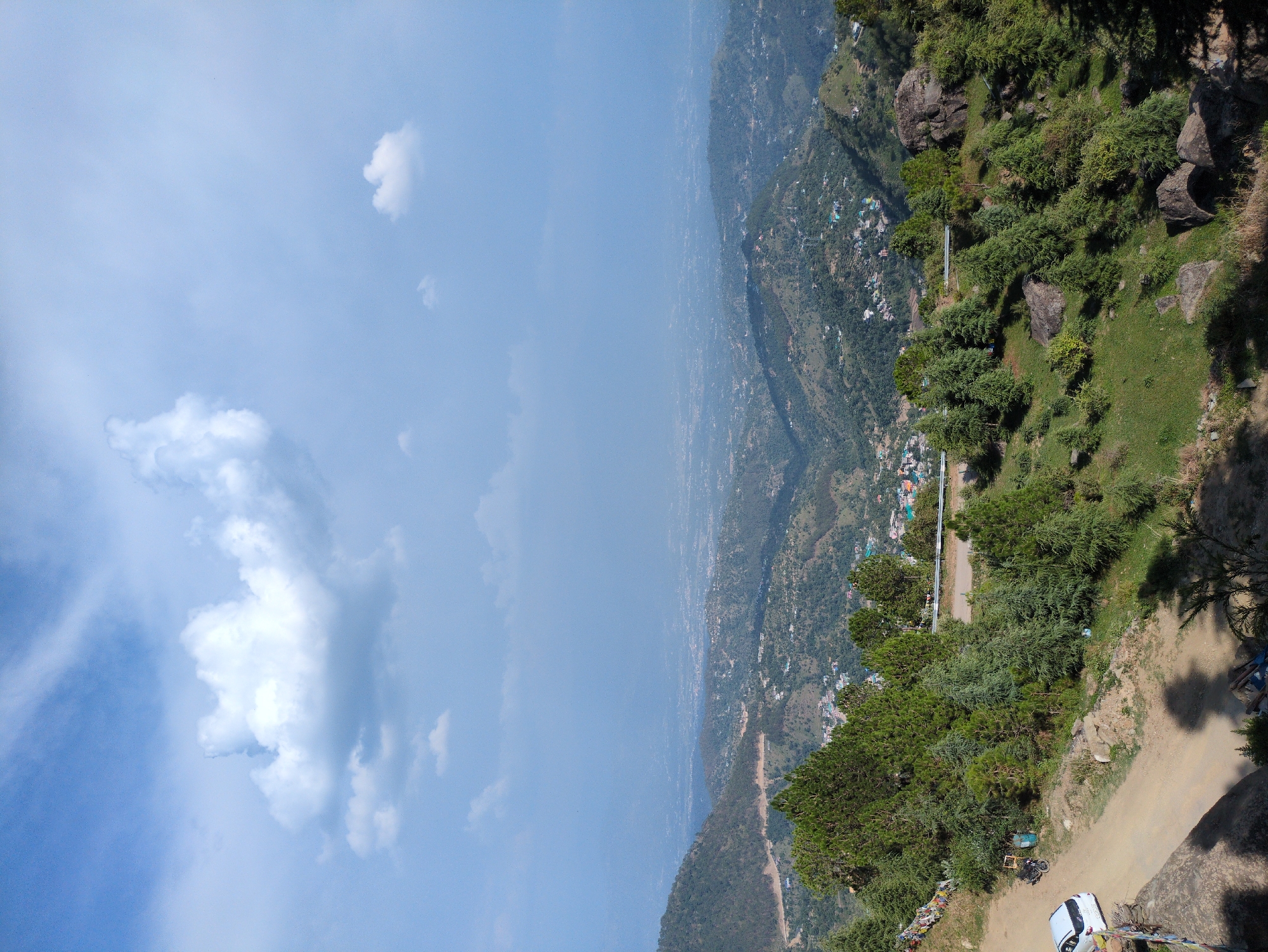 An elevated, wide-angle shot captures a verdant, mountainous landscape under a partly cloudy sky. In the foreground, a dirt road curves along the edge of a hillside, with a white car parked on the left and a motorcycle and trash receptacle further up the road. Colorful prayer flags are strung along a stone wall to the left of the road. The hillside is covered in lush green grass and scattered rocks, dotted with numerous coniferous trees of varying sizes. In the midground, more rolling hills are covered in trees, with a small town visible nestled in the valleys between them. The background shows a vast expanse of hazy, distant hills and a low-lying city spread out in the valley, suggesting the city of Doh, India. The sky is a light blue with large, fluffy white clouds, some casting shadows on the terrain below.