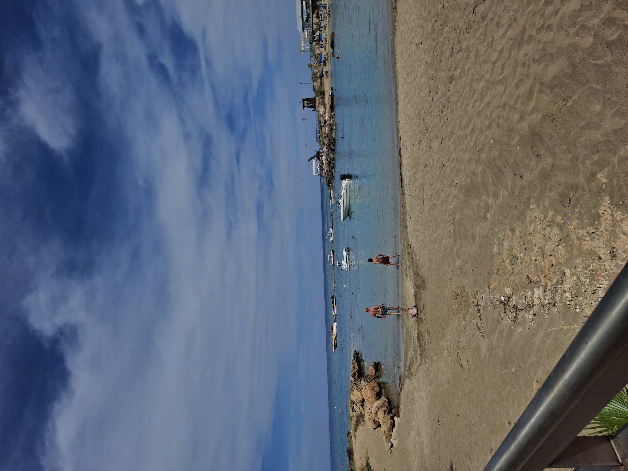 The image depicts a beach scene at Fig Tree Bay in Protaras, Cyprus. Two individuals are wading in the shallow turquoise water. Several boats are anchored further out in the bay. The beach is sandy, with some rocky outcrops along the shoreline. A lifeguard station is visible in the distance. The sky is blue with scattered clouds. A railing and vegetation are partially visible in the foreground.