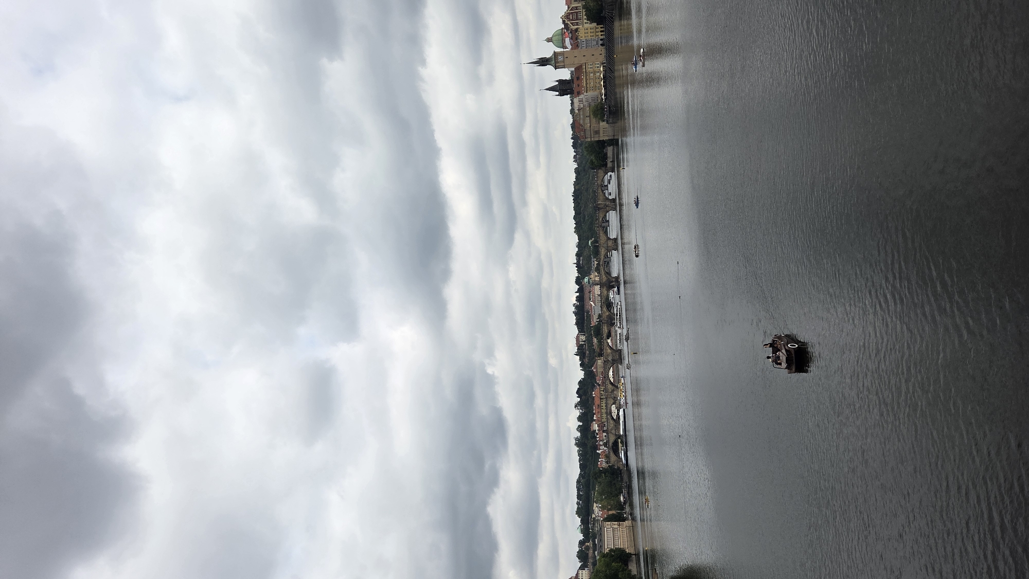 A wide shot captures the Vltava River in Prague, Czechia, under an overcast sky filled with thick, gray clouds. The river dominates the foreground, its surface a dark, reflective expanse. In the mid-ground, a small, dark-colored boat with two people onboard navigates the water. Further back, the iconic Charles Bridge stretches across the river, supported by numerous arches. Buildings with red-tiled roofs line the riverbanks, interspersed with greenery and architectural structures, including towers and spires. Several other smaller boats and what appear to be swans or ducks are scattered across the river and in the distance. The overall scene depicts a moody, atmospheric day in the historic city.