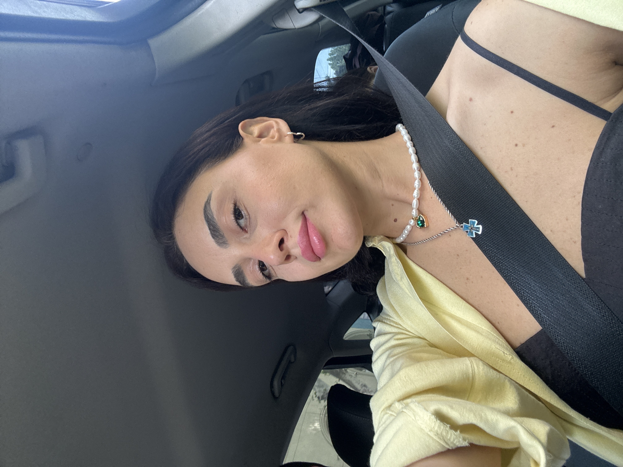 A medium shot selfie of a woman in the passenger seat of a car, likely in Tbilisi, Georgia, captures her from the chest up. She is wearing a light yellow open button-down shirt over a black tank top, accessorized with a pearl necklace featuring a green pendant and a silver necklace with a blue cross pendant. A black seatbelt crosses her chest. Her dark hair is down, and she has distinct eyebrows, prominent cheekbones, and full lips. The background shows the car's interior, including the ceiling, window frame, and a portion of a car seat. The lighting suggests it is daytime, with natural light filtering in from the side.
