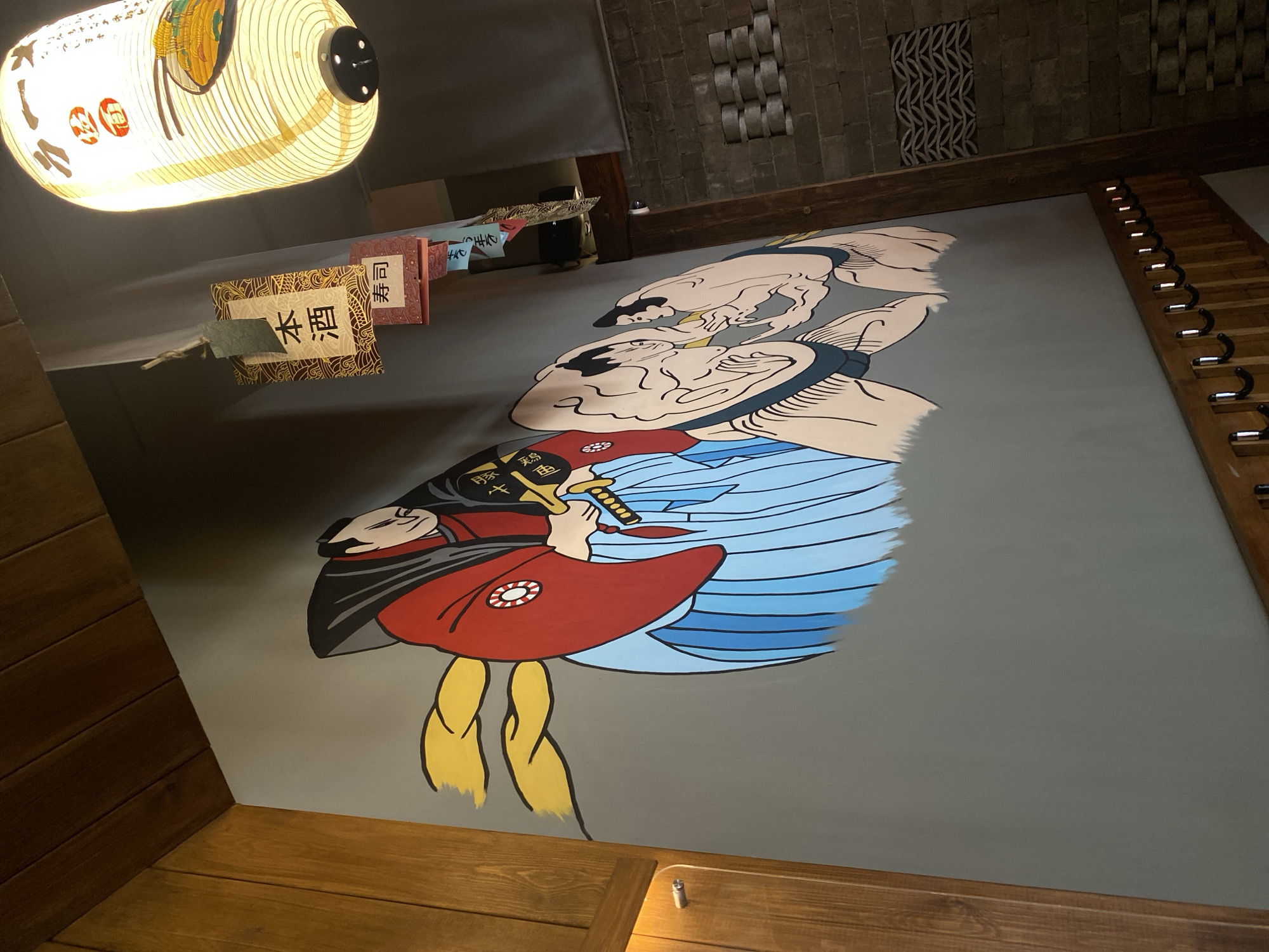 The image shows an interior scene in what appears to be a restaurant. A large wall mural dominates the view, depicting two figures in a Japanese-style art. The upper figures appear to be sumo wrestlers. The lower figure is holding a sword. Above the mural hangs a traditional Japanese paper lantern. To the left of the mural are hanging paper menus with Japanese writing. The floor is wooden. There are hooks in the upper right corner of the image. The location in the city of Krasnoyarsk, Russia is not indicated.