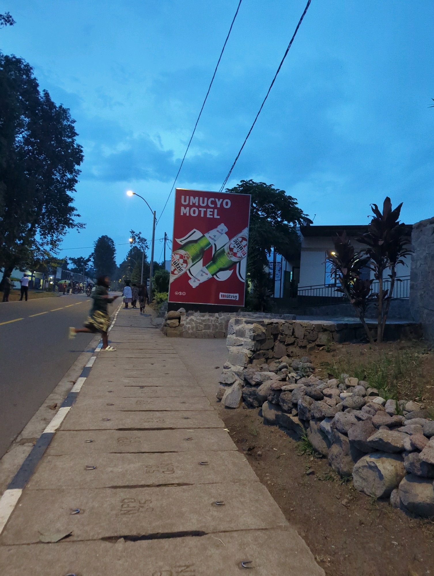 The image depicts a street scene at dusk in Musanze District, Rwanda. A large red billboard advertising "UMUCYO MOTEL" with images of beer bottles is prominently displayed on the right side of the frame. A sidewalk runs along the street, with several individuals walking on it. In the foreground, a person in motion is captured on the sidewalk, suggesting activity and movement. The street itself appears to be paved, with a white and black striped curb. The environment is characterized by a dusky sky with a light blue hue, indicating the transition from day to night. Trees and some buildings are visible in the background, suggesting an urban or semi-urban setting. The overall scene conveys a sense of everyday life in the Musanze District as the day winds down.
