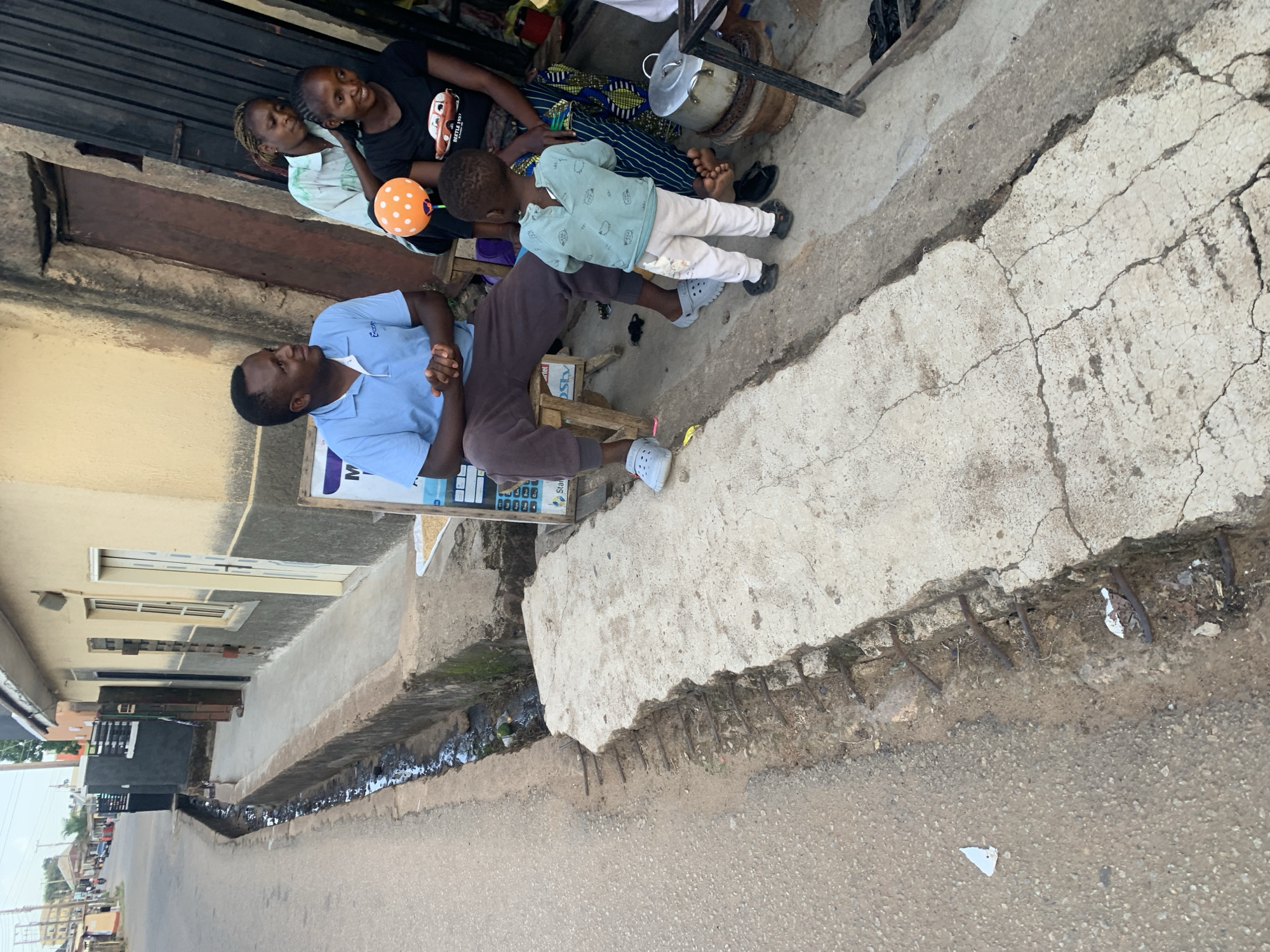 In Jos, Nigeria, the image captures a street scene. A man in a light blue shirt and brown pants sits on a wooden stool. Nearby, two women sit; one wears a black shirt, and the other wears a light-colored shirt. A child stands beside them, dressed in a light-colored top and pants. An orange balloon is also visible. They are positioned on a concrete surface beside a building. The foreground features a street with a drainage ditch running along the edge. The concrete curb along the street shows exposed rebar and cracks.