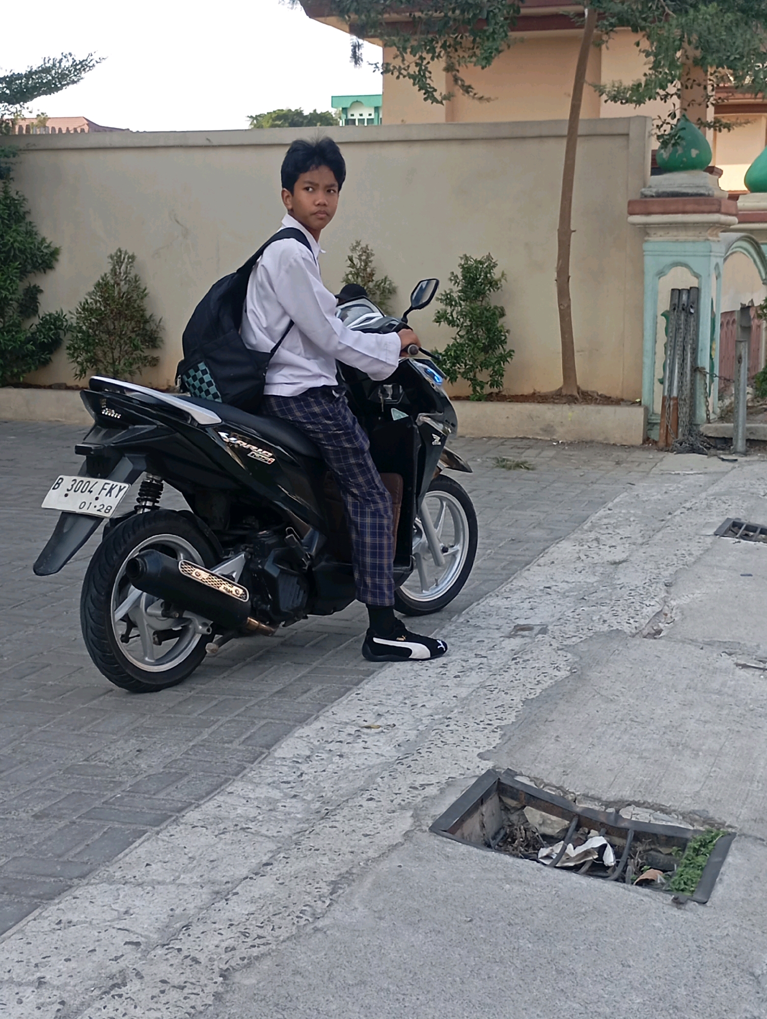 A young boy wearing a white shirt, plaid pants, and black sneakers with white accents sits on a black motorcycle. He has a black backpack on his back. The motorcycle is parked on a paved surface. In the background, there is a beige wall, some green bushes, and a building with a green dome. A drain grate is visible in the foreground. The scene appears to be in Babelan, Indonesia, given the context of the user's request.
