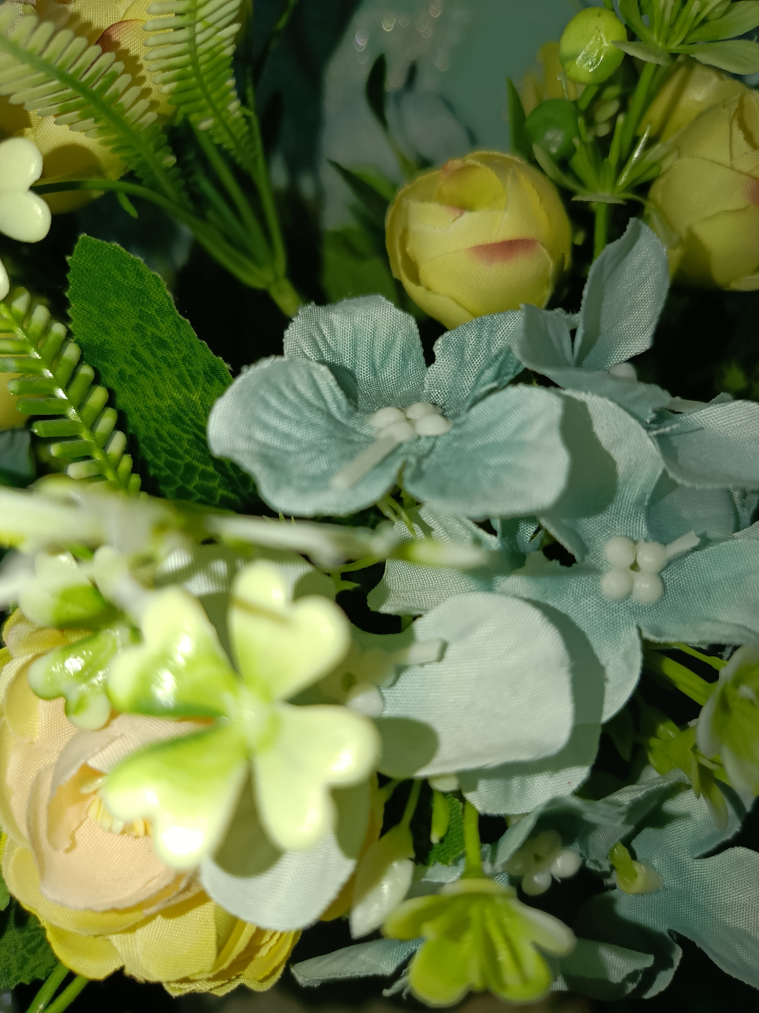 The image is a close-up, macro shot of a bouquet of artificial flowers. The bouquet features several types of flowers, including light yellow roses, light blue hydrangeas, and small green leafy accents. The lighting appears to be artificial and is creating highlights and shadows on the petals. The background is dark and out of focus, emphasizing the floral arrangement. There is no indication of a specific location in Tashkent, Uzbekistan, nor are there any people or actions depicted.
