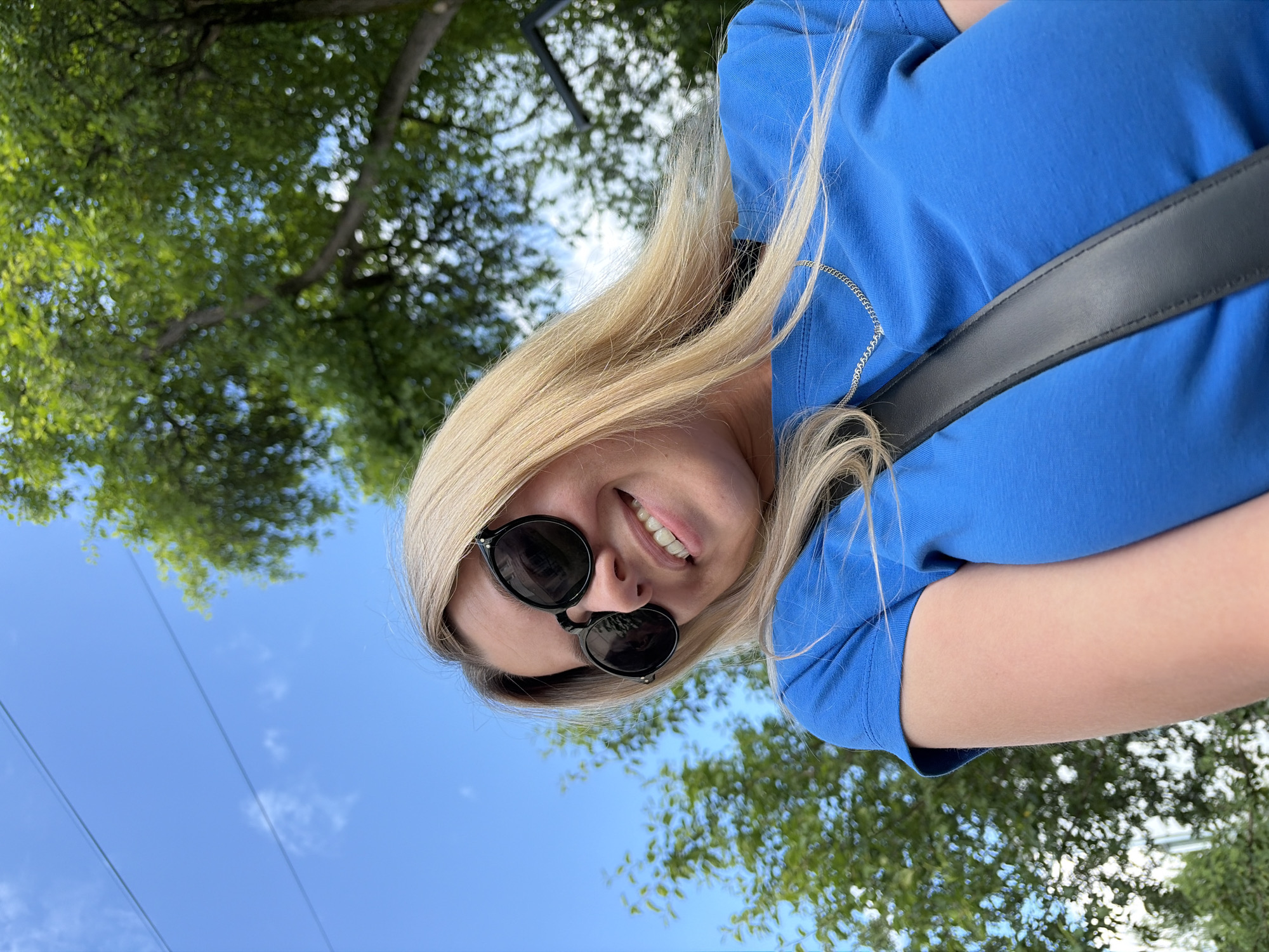 A blonde woman wearing sunglasses and a blue t-shirt is photographed from a low angle outdoors. She is smiling and looking down, with her hair falling forward. Trees and a blue sky with some clouds are visible in the background. The setting appears to be a park or outdoor public space. The image does not provide any specific clues to identify its location in the city of Chaikovsky, Russia.