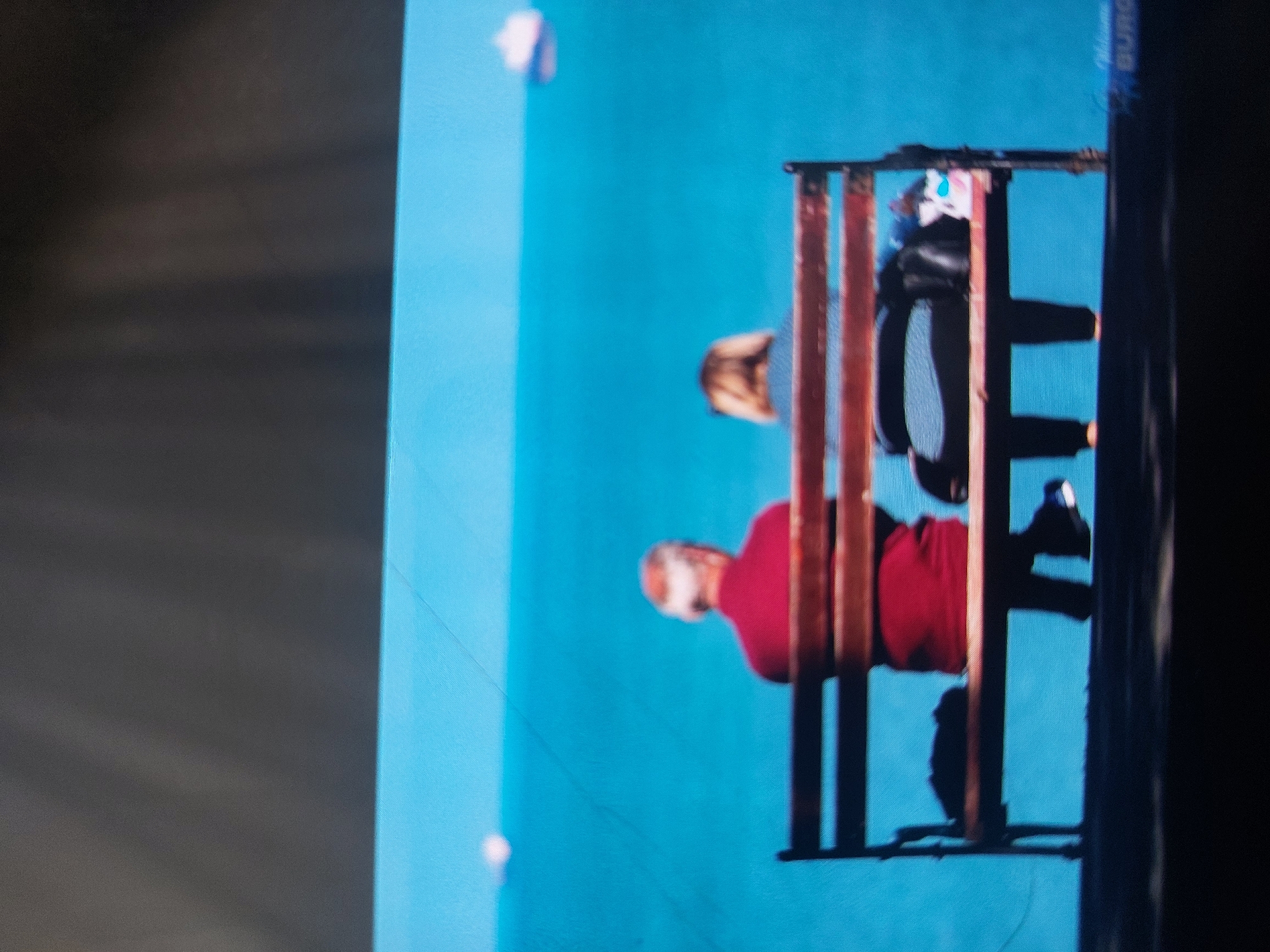 The image shows a scene in Tsarevo, Bulgaria. Two people are sitting on a wooden bench. A man is wearing a red shirt. A woman is wearing a striped shirt and black pants. The background is a bright blue color. A white object is visible in the background.