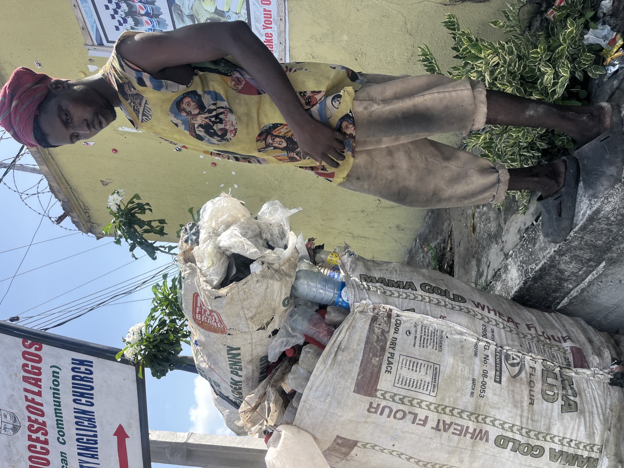 The image captures a scene at a street corner in Ado, Nigeria. A man stands beside a pile of discarded materials packed in sacks labeled "Mama Gold Wheat Flour". He wears a yellow t-shirt with face images and trousers. A sign is visible, showing the name "Diocese of Lagos, Holy Anglican Church". White flower with green leaves are also present. The environment appears to be urban, with visible walls, posters, and vegetation.