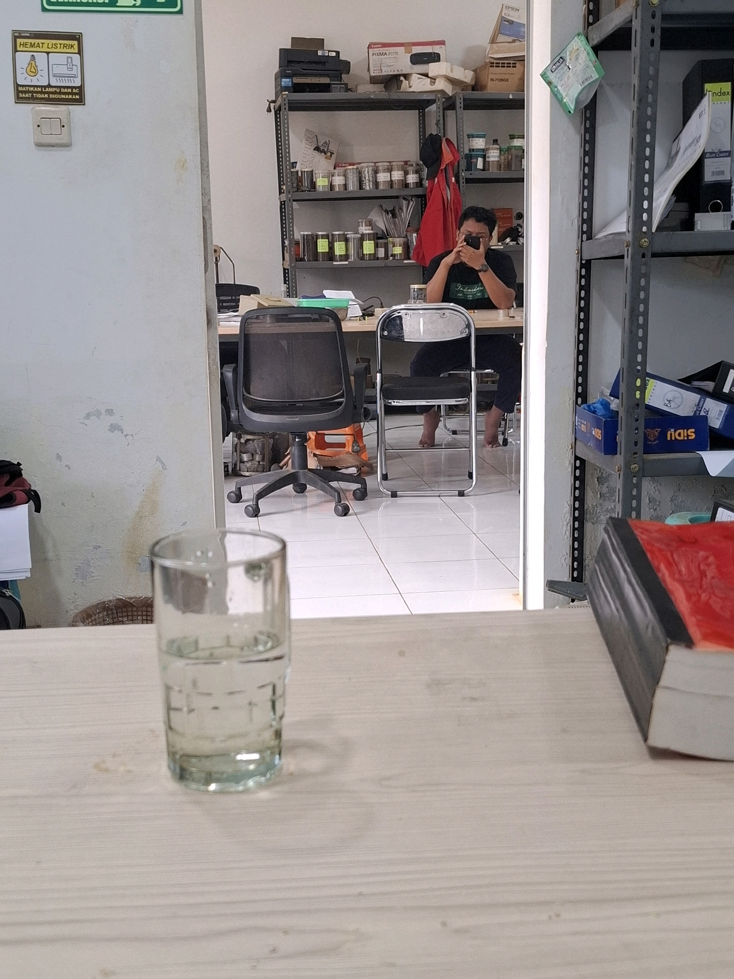 The image depicts an indoor scene, likely an office or storage area, with a focus on a glass of water in the foreground. The scene is viewed from a low angle, looking across a light-colored wooden surface. Behind the glass, a white door is partially open, revealing a room with shelves filled with various items, including jars, boxes, and binders. A person is visible in the reflection of the door, sitting at a desk and holding a camera to their face, seemingly taking a picture of themselves. Two chairs are present in the room, one a black office chair with wheels, and the other a folding chair. The environment appears somewhat cluttered, with stacks of paper and office supplies visible on shelves and desks. The lighting is neutral, suggesting an indoor setting. The text on the sign near the door indicates "HEMAT LISTRIK" which translates to "SAVE ELECTRICITY" in Indonesian, along with instructions to "MATIKAN LAMPU DAN AC SAAT TIDAK DIGUNAKAN," meaning "TURN OFF LIGHTS AND AC WHEN NOT IN USE." This suggests the location is in Indonesia, as the text is in Bahasa Indonesia. While a specific city is not explicitly mentioned in the image itself, if it were located in Kutasirna, Indonesia, the scene would represent a typical office or utility space within that region.