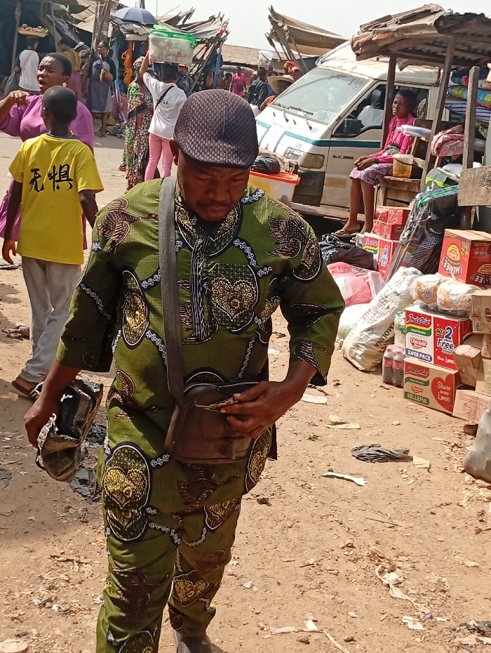 The image captures a bustling outdoor market scene, likely in Onitsha, Nigeria, under bright daylight conditions with strong overhead sunlight casting shadows. The main subject is a man in the foreground, wearing a distinctive olive green patterned tunic and matching trousers, along with a dark, textured cap. He carries a dark brown leather shoulder satchel and holds a pair of dark shoes in his left hand while looking down, seemingly engaged with his satchel or counting something.
In the mid-ground, a woman in a purple top walks to the left, and beside her, a child in a yellow t-shirt walks away from the camera. The back of the child's t-shirt displays the Chinese characters "無惧" (Wú Jù), which translates to "Fearless." Other market-goers are visible further back, including a woman balancing items on her head under a dark umbrella.
To the right, a white passenger van is parked, with a woman seated in its open side door. The surrounding area is filled with makeshift market stalls constructed from wood and corrugated metal, displaying various goods. The ground is unpaved dirt. Merchandise includes numerous stacked cardboard boxes of instant noodles such as "Indomie" and "NIMMEE," as well as "Super Pack" items, "MOLINA" products, and "COCA-COLA" bottles. Some boxes also show text like "FREE FROM ARTIFICIAL FLAVOURS & COLOURS," "BELLEFUL," and "Show Your Love." The overall atmosphere is busy, typical of an active open-air market.