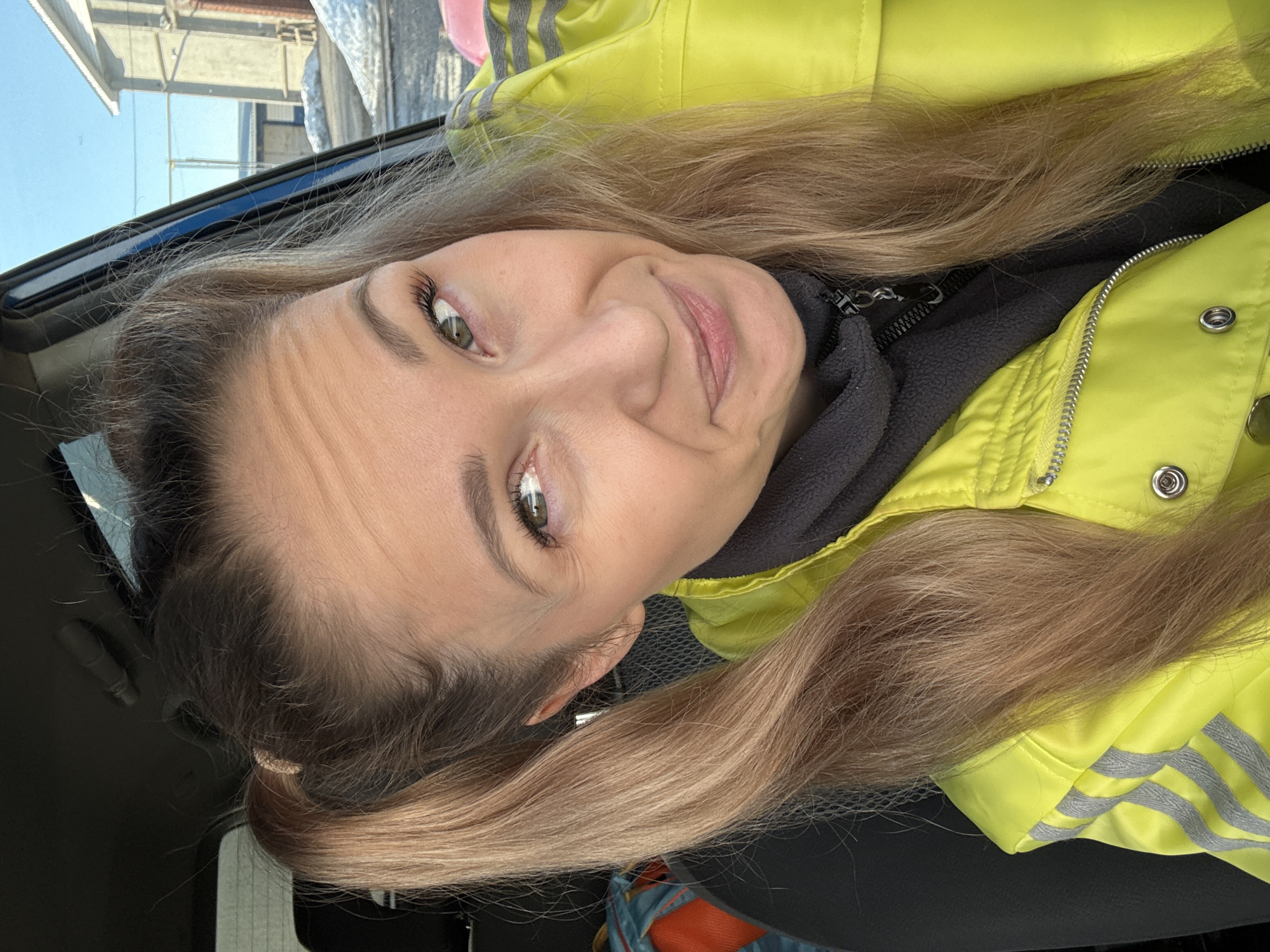 A woman with fair skin, long blonde hair pulled back into two low pigtails, and green eyes is looking directly at the camera and smiling slightly. She is wearing a bright yellow jacket with three grey stripes on the sleeves. She is wearing a grey sweater under the jacket. The photo appears to have been taken inside a car. The car window is visible in the background, showing a partially cloudy blue sky and blurred buildings. There are also snow and ice formations visible.
The image does not provide sufficient information to ascertain the location within the city of Chaikovsky.