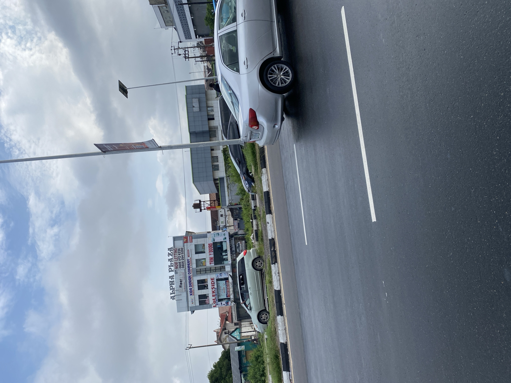 This image depicts a street scene in Ojomu, Nigeria, on an overcast day. The foreground shows a dark asphalt road with two solid white lane markings. The road surface appears wet or reflective. A raised median strip, edged with a black and white striped curb, separates the traffic lanes.
On the median, a light green minivan, identifiable as a Toyota Sienna, is parked facing left. Behind it, a dark-colored sedan, possibly a Hyundai Elantra, is also parked, partially obscured. A single individual wearing dark clothing is visible near the rear of the dark sedan, moving away from the viewer.
Dominating the background on the left is a two-story building named "ALPHA PLAZA," prominently displayed in large white letters. Below the plaza name, a sign reads "NOW LETTING" followed by two phone numbers: "0806 412 4226" and "0806 284 5290." The first floor features a business called "KIDS DESIGN COMPANY." The ground floor displays "LaLEKSIDE" and "TRINI Y DOORS" as storefronts. Adjacent to Alpha Plaza, to the left, are older buildings with reddish roofs amidst green foliage.
Further down the road, to the right of Alpha Plaza, a modern building with dark grey or blue cladding is visible. A tall utility pole with multiple cross-arms and power lines stands near Alpha Plaza. In the mid-ground, a silver Toyota Corolla sedan is observed on the road, partially facing left. Its rear bumper and side profile are visible, including its alloy wheels.
To the right of the frame, a tall, slender metal pole features a dark red or maroon banner, mostly unreadable. Another similar pole, topped with a solar panel, indicates a street light. The sky is heavily clouded with a mix of grey and white, suggesting an overcast day with no direct sunlight.