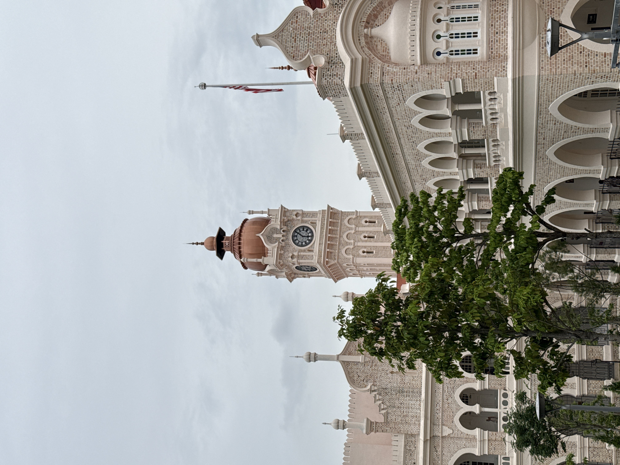 The image shows the Sultan Abdul Samad Building in Kuala Lumpur, Malaysia. The building's architecture includes a clock tower, arched windows, and decorative brickwork. A Malaysian flag flies atop one of the building's spires. Part of a tree with green leaves is visible in the foreground. The sky is overcast.