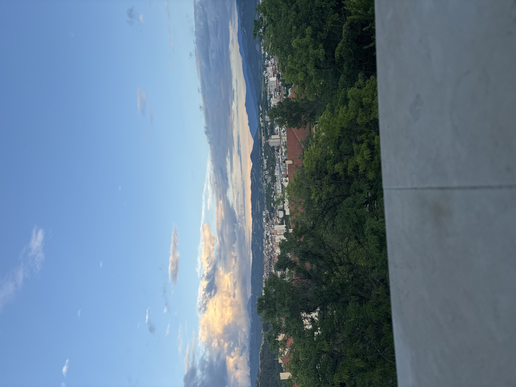 A panoramic outdoor photograph captures a late afternoon or early evening view of Blagoevgrad, Bulgaria, from an elevated vantage point.
The upper portion of the frame is dominated by a vast sky, transitioning from clear blue on the left to a more dramatic display of clouds on the right. These clouds vary from sparse, wispy white formations to larger, more substantial cumulus clouds, tinged with warm hues of orange, pink, and purple, indicating the approach or presence of sunset.
Below the sky, a distant mountain range stretches across the horizon, appearing hazy and blueish-grey. Nestled in the valley between the viewer and the mountains is the city of Blagoevgrad, with numerous buildings and structures densely packed together. Various architectural styles are visible, including residential and commercial buildings, some with distinctive red-tiled roofs.
The foreground on the right side of the image features a dense canopy of lush green trees, suggesting a park or forested area on a hillside. Partially visible on the far right is a light-colored, possibly concrete or stone, wall or railing, indicating the photo was taken from a balcony, terrace, or observation deck.
No people or discernible text are visible in the image. The overall impression is one of serene natural beauty encompassing an urban landscape, bathed in the soft, warm light of dusk.