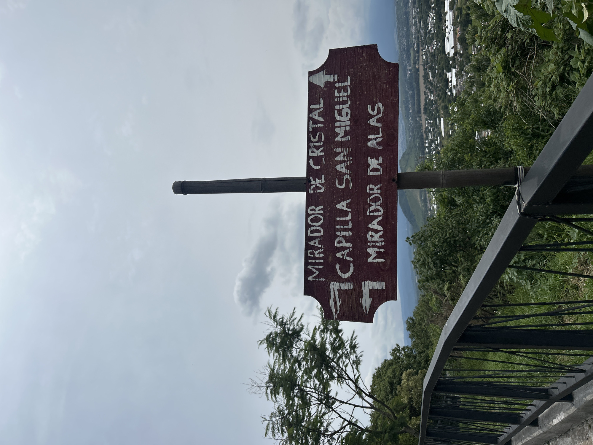A wooden signpost with three directional signs is positioned against a cloudy sky, overlooking a city in the distance. The signpost, made of a dark pole, has three maroon-colored signs with white lettering. The top sign reads "MIRADOR DE CRISTAL" with an arrow pointing right. The middle sign says "CAPILLA SAN MIGUEL" with an arrow pointing left. The bottom sign reads "MIRADOR DE ALAS" with an arrow pointing left. To the left of the signpost, a leafy green tree is visible. In the foreground, a black metal railing runs horizontally across the frame, with a concrete walkway beneath it. Beyond the railing, dense green foliage covers the hillside. The city in the background appears to be Atlixco, Mexico, with buildings and structures spread across the landscape, nestled among rolling hills. The overall scene suggests an elevated viewpoint in a semi-urban, natural setting.