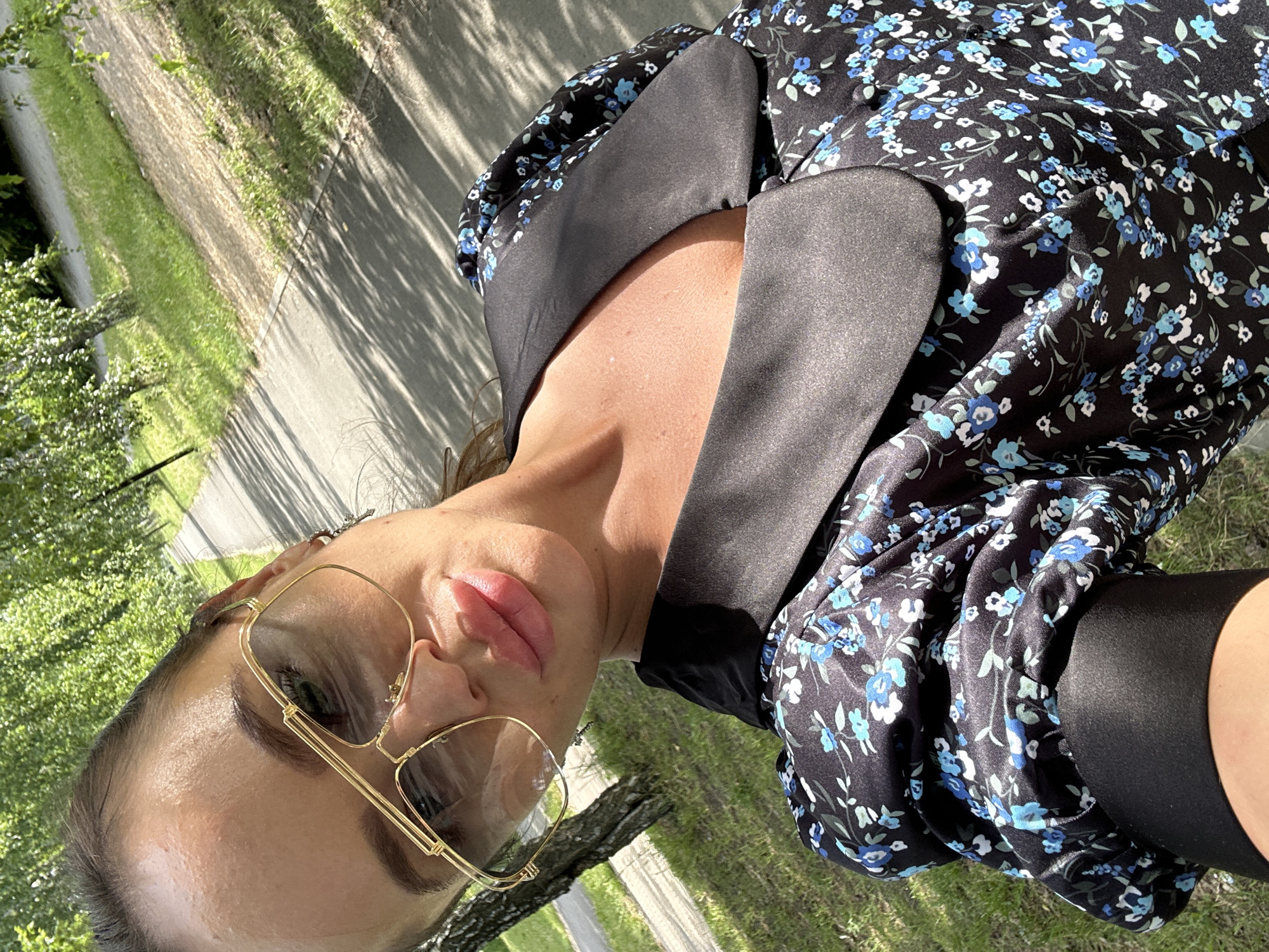 This is a vertical, medium shot photograph of a woman wearing a black floral dress and gold-rimmed aviator sunglasses. The woman is outdoors, with a path and grassy area visible in the background, suggesting a park or a street with greenery. Sunlight casts shadows from trees onto the path, indicating it is daytime, likely afternoon due to the length and angle of the shadows. The woman's face is partially obscured by her sunglasses, but her lips are slightly parted, and her skin appears bronzed. She is wearing dark leggings or tights on her legs. The floral pattern on her dress consists of small blue and white flowers with green leaves scattered across a black background. The overall mood is casual and relaxed, with the natural lighting and outdoor setting contributing to a sense of warmth. The image appears to be a selfie, taken by the woman herself.