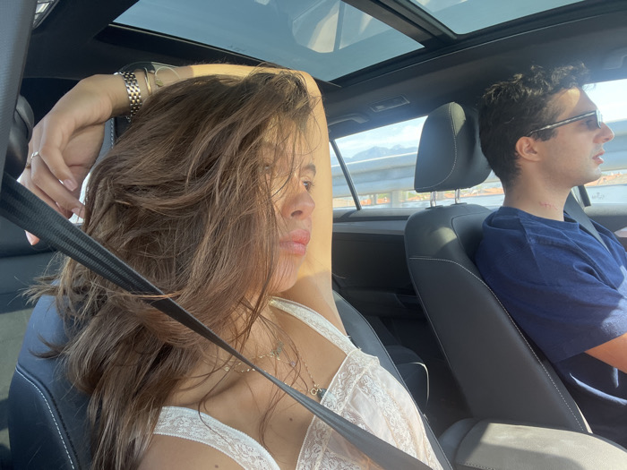 Inside a vehicle, likely a car, two individuals are present during daytime in Camaiore, Italy. In the foreground, a female passenger with long brown hair, wearing a light-colored, possibly lace, top, is positioned in the front passenger seat. Her right arm is raised, with her hand resting behind her head, and a wristwatch is visible on her left wrist along with a ring on her left ring finger. A necklace with multiple small charms is visible around her neck. A black seatbelt crosses diagonally over her chest. Her face is partially obscured by her hair and angled towards the right side of the frame. In the background, a male driver, wearing sunglasses and a dark blue t-shirt, is visible in profile, looking forward. Red marks are present on the left side of his neck. The vehicle features a panoramic sunroof and black leather seats. Through the windows, a bright, sunny outdoor environment is visible, including a road barrier and distant mountains under a clear sky.