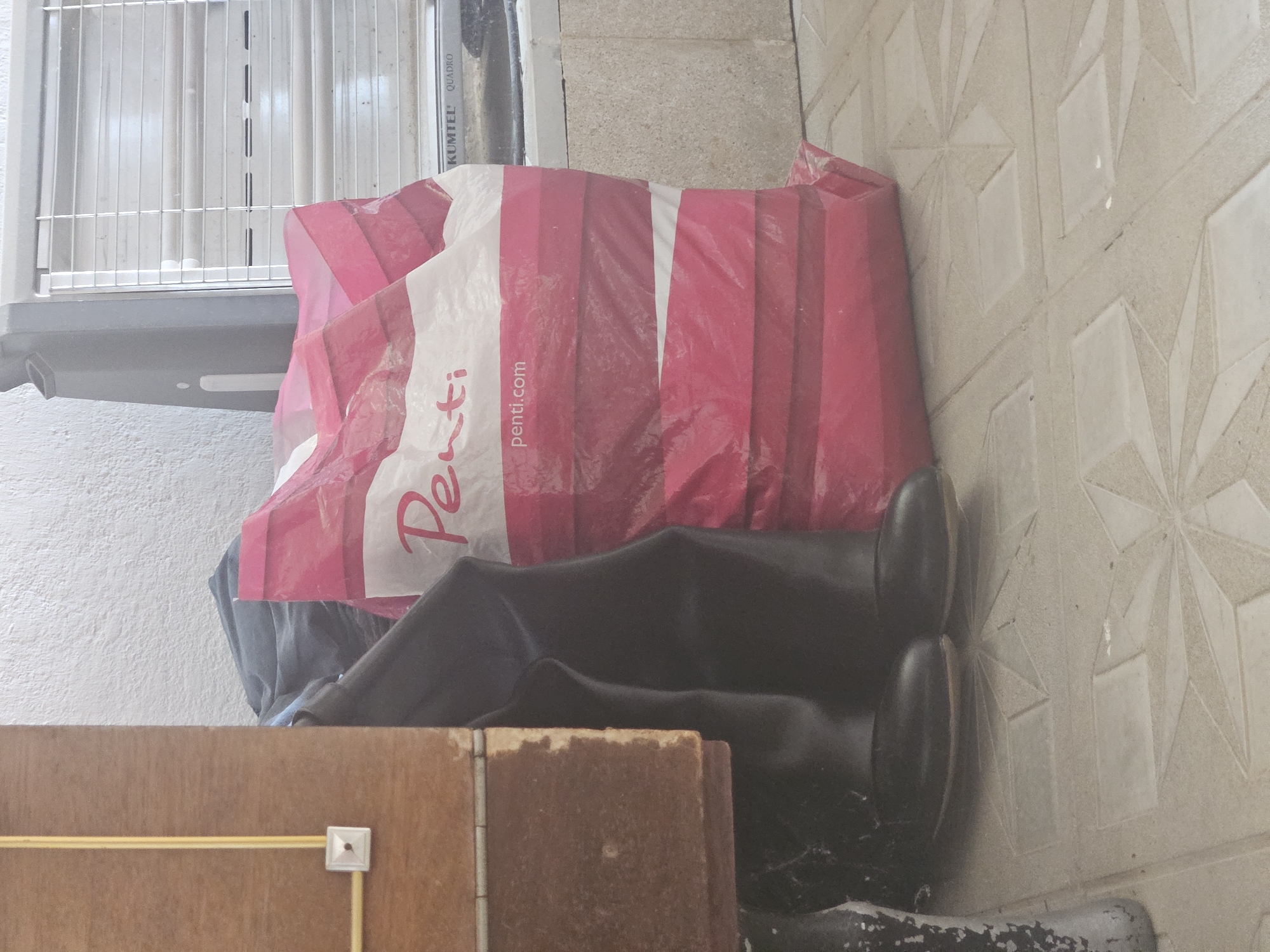 A high-angle shot shows a pair of dark boots on a tiled floor. To the left of the boots is a wooden structure with a light yellow electrical conduit and a white outlet. To the right of the boots is a large, pink and white striped plastic bag. Behind the bag is a light gray electric heater with horizontal grates. The wall behind the objects is textured and white.