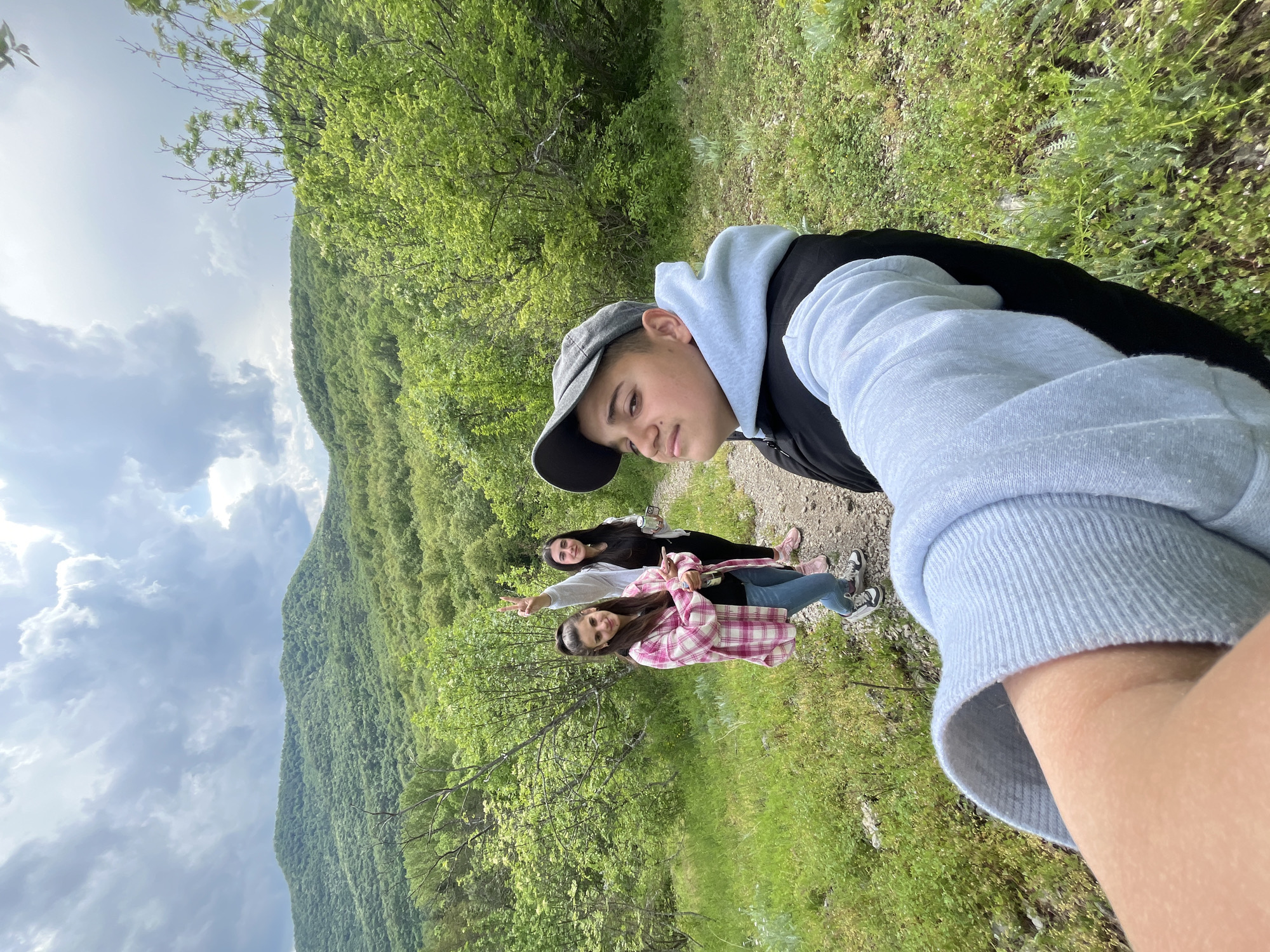 A group of three young people are on a trail in Kamchia, Bulgaria. The foreground features a young man taking a selfie. He is wearing a backward gray cap, a black jacket, and a light blue hooded sweatshirt. Behind him, two young women are walking together. One woman is wearing a pink and white plaid shirt and the other is wearing a gray sweatshirt. The background consists of a green mountainside and a cloudy sky. The trail is surrounded by green grass and trees.