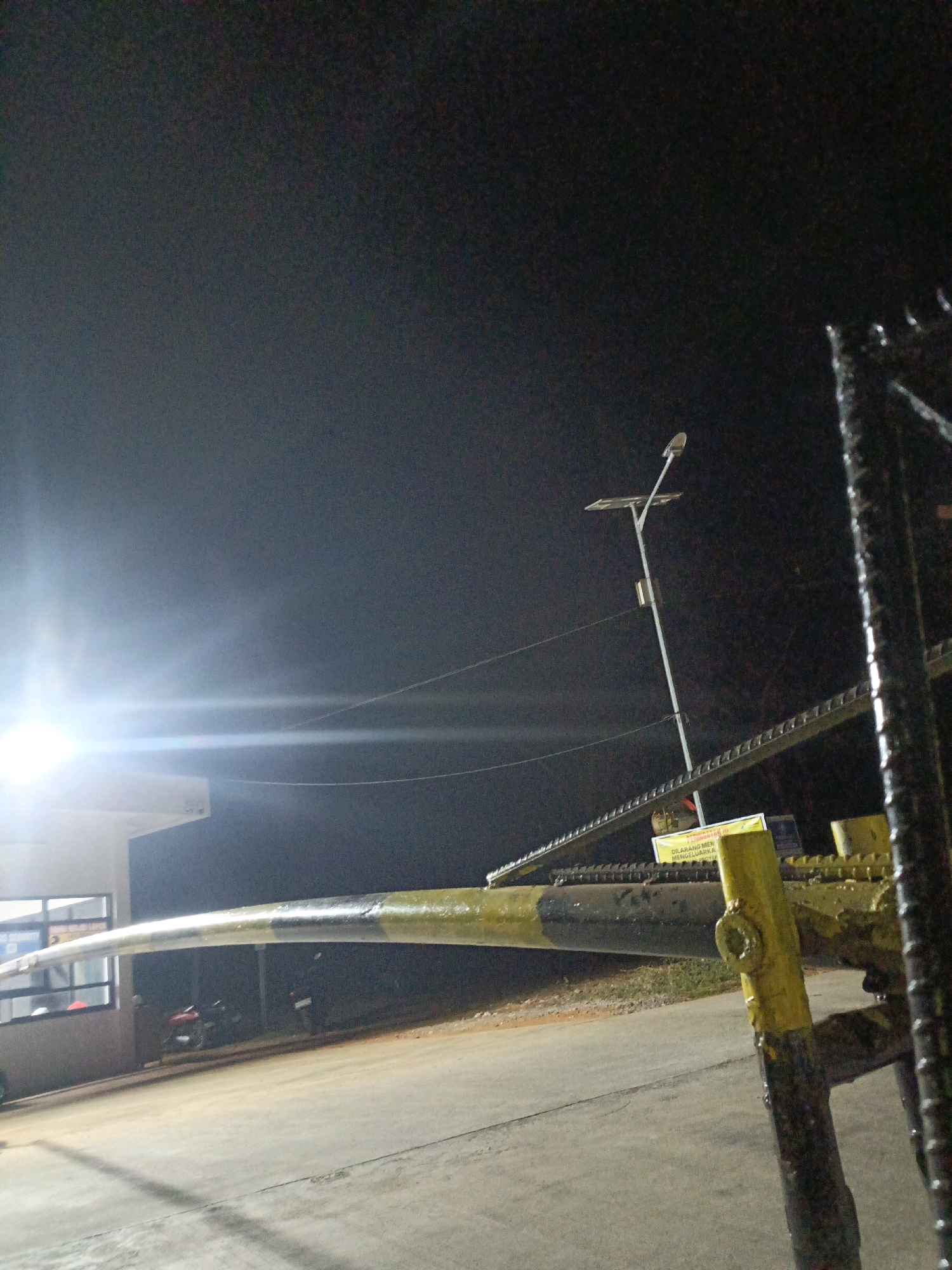 The image depicts a barrier arm at what appears to be an entrance or checkpoint in Jenu, Indonesia. The barrier arm, painted yellow and black, is lowered across a paved road. Behind the barrier arm, a building with windows is visible, possibly a guardhouse or administrative office. A street light with solar panels stands tall in the background, illuminated against the dark night sky. Several motorcycles are parked near the building. The scene is lit by artificial lights, creating strong reflections and a slightly hazy atmosphere. The overall impression is that of a secure access point, likely operational at night.