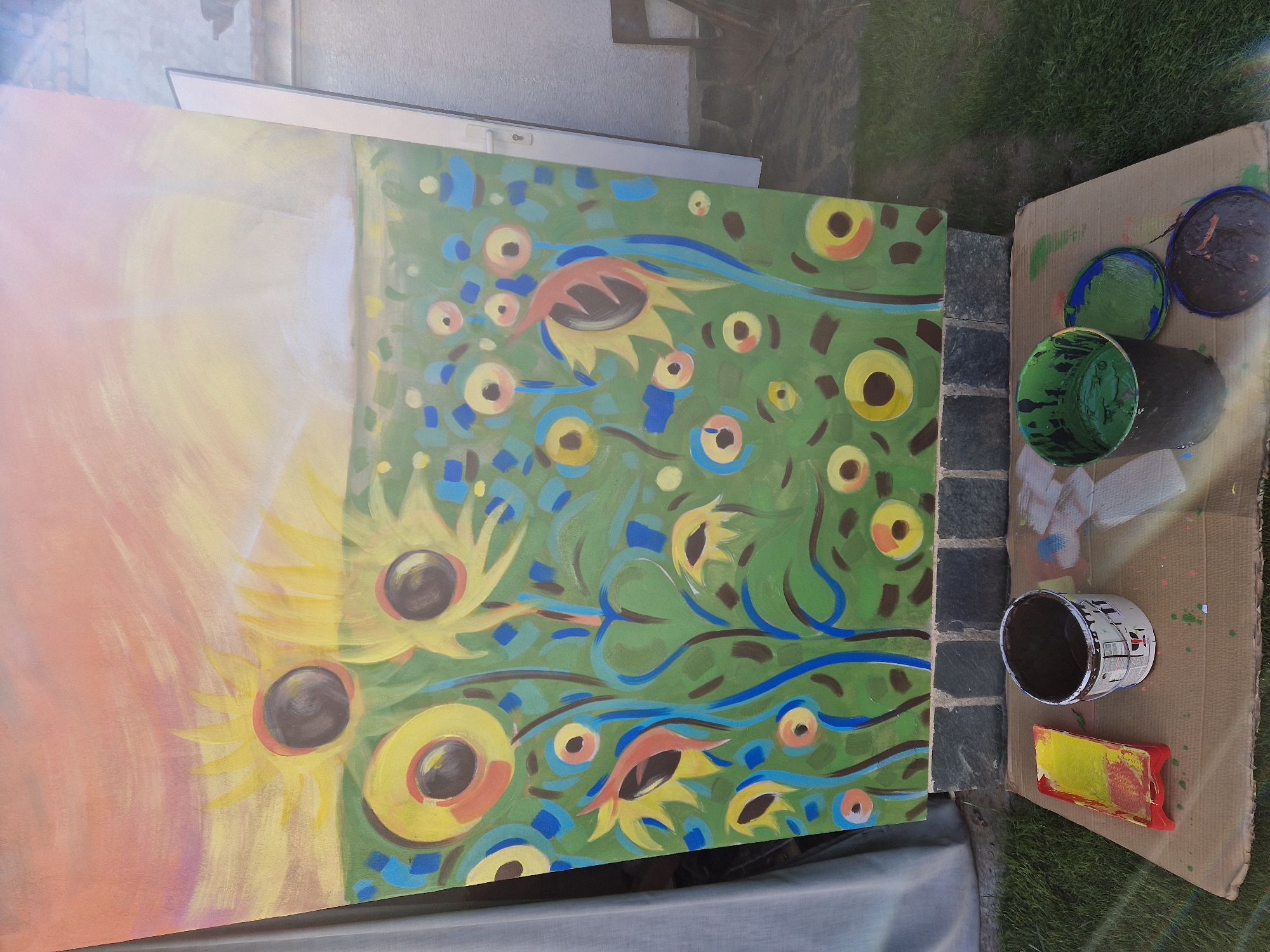 The image shows a painting in progress, seemingly outdoors, with two adjacent canvases. The left canvas features a sunny sky with diffused colors transitioning from orange to yellow. The right canvas displays a vibrant, stylized sunflower field with green as the dominant background color. The sunflowers are depicted with bold, geometric shapes and colorful accents like blue and brown. Nearby, there's a piece of cardboard used as a painting surface, containing paint cans and color mixing trays. The environment consists of bricks and grass.