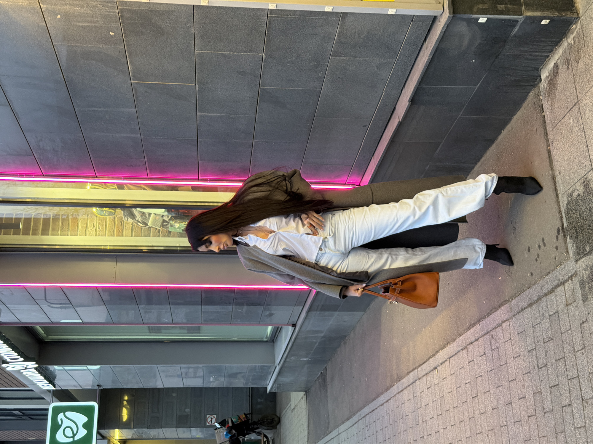 The image depicts a single female subject standing on a city sidewalk in front of a building facade. The location is Bolshaya Pirogovskaya Ulitsa, 13, building 2, Moscow, Russia, identified by a green sign reading "МЕРКУРОВА СТОМАТОЛОГИЯ" (Merkurova Dentistry) partially visible on the building to the left.
The female subject, possessing long dark hair, is dressed in a white collared shirt, light-wash denim pants, a long dark grey coat, and black pointed-toe ankle boots. She holds a tan leather handbag with a gold-colored clasp in her left hand, which rests at her side. Her right hand is placed on her hip, partially covered by her coat. Her posture indicates she is posing, with her body facing generally forward and her head turned to her right, looking away from the camera.
The building behind her features a dark grey tiled facade. To the subject's left, a "No Smoking" sign is visible on the wall above a dark-colored scooter or moped parked near the curb. A large window display to her right is accentuated by vertical pink neon LED strip lights along its edges, and warm yellow light emanates from within the display. The sidewalk beneath her consists of a concrete slab with a visible seam separating it from a section of light-colored brick paving to the left. The scene is illuminated by a combination of ambient daylight and the artificial neon and interior lights.