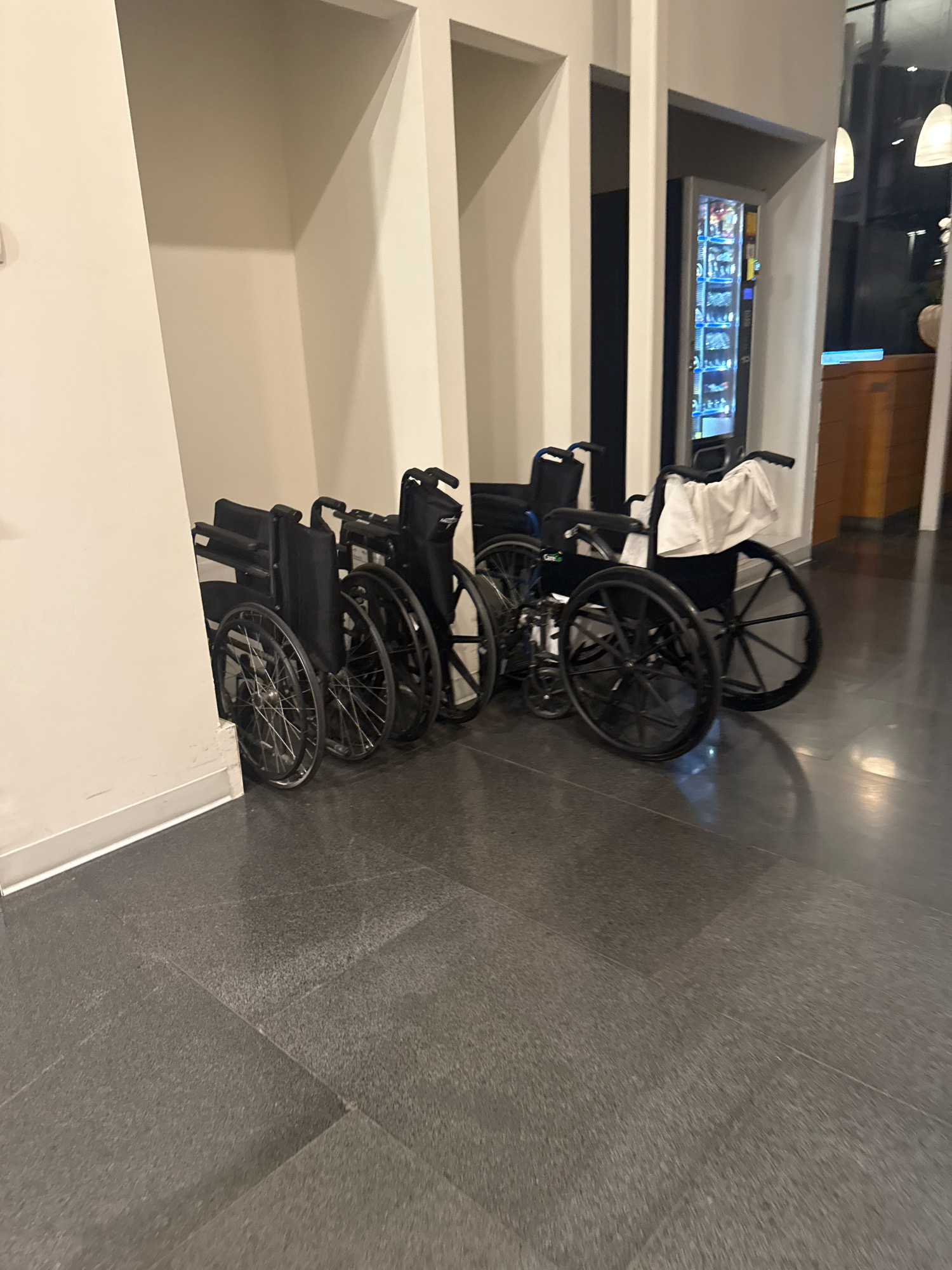 In a building with beige walls and dark tiled flooring, several wheelchairs are parked against a wall. To the right of the wheelchairs, a vending machine with illuminated shelves is visible. The scene appears to be indoors, possibly in a hospital or public facility, with no people present. The lighting is dim, with artificial lights casting reflections on the floor.