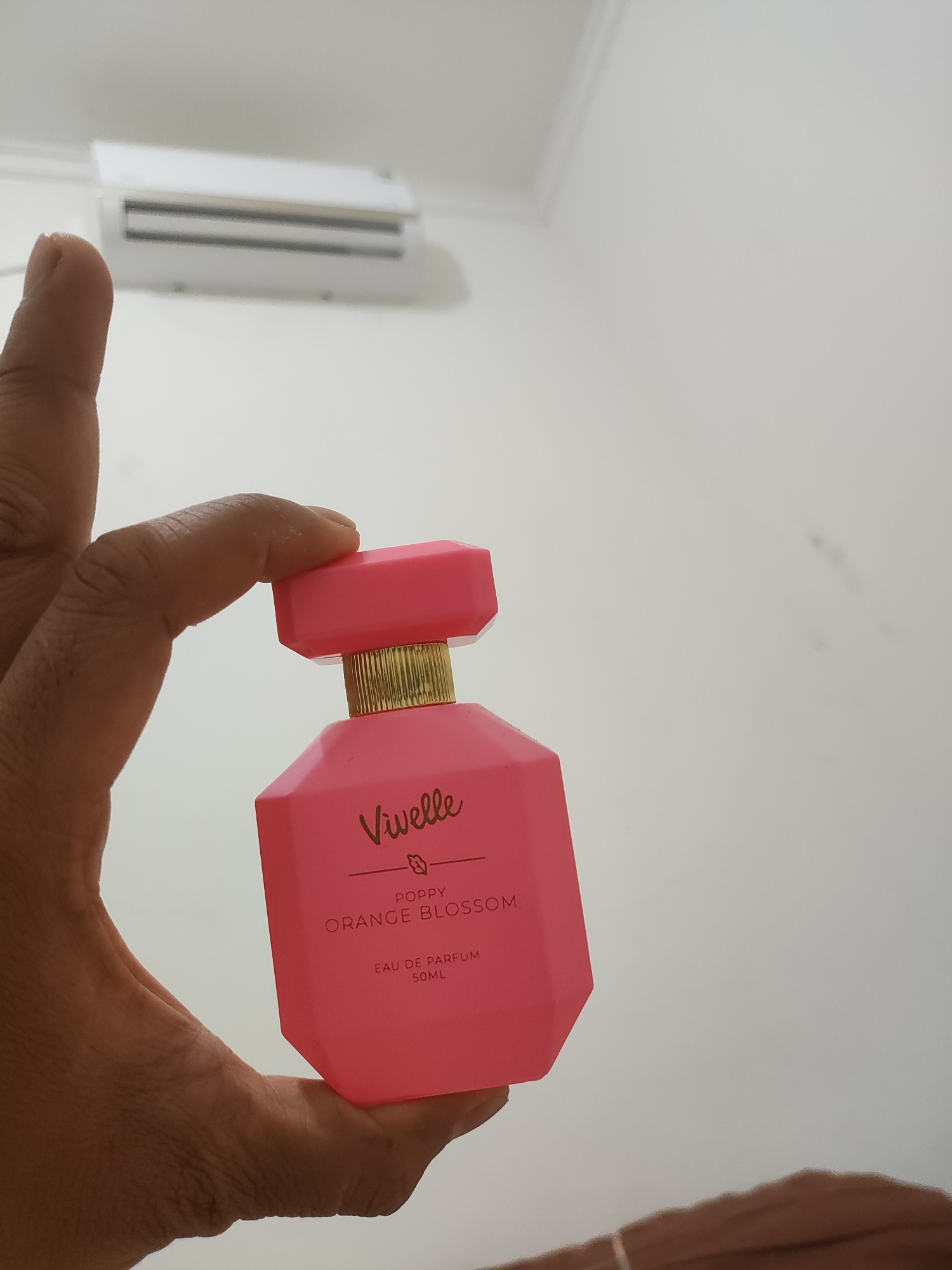 A person's thumb and index finger hold a bright pink, faceted perfume bottle with a gold band around the neck. The bottle has text printed in a darker pink, reading "Vivelle", a small leaf icon, "POPPY ORANGE BLOSSOM", "EAU DE PARFUM", and "50ML". The background is a plain white wall with a white air conditioning unit mounted high on it. The scene appears to be indoors.