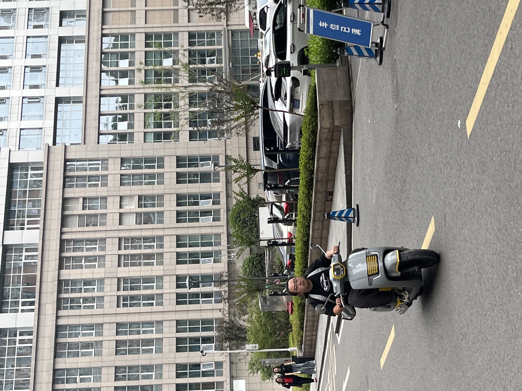 In Yuzhong District, China, a man wearing glasses and a black t-shirt is riding a gray and yellow electric scooter. He is on an asphalt road with yellow lane markings. Behind him, a modern building with many windows stands tall. The building's facade is made of light-colored stone. Several cars are parked on the side of the road, and there are some trees and bushes in the background. A blue and white sign with Chinese characters is visible on the right side of the image.