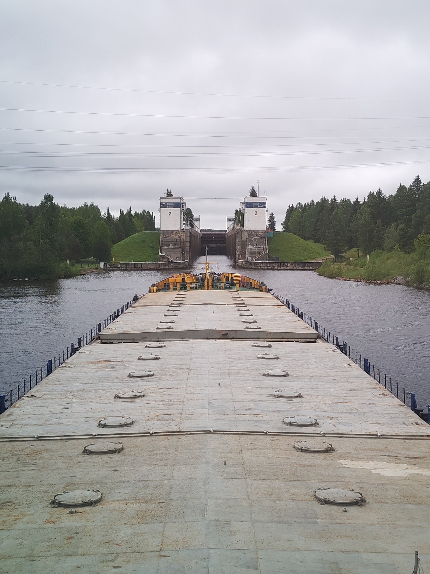 The image shows a view from the deck of a barge moving toward Lock Number 2 of the Volga-Baltic Waterway, located in Анхимово, Russia. The lock structure is visible in the distance, flanked by green, forested banks. The deck of the barge is concrete and features multiple circular hatches. The sky is overcast. Power lines stretch across the sky.