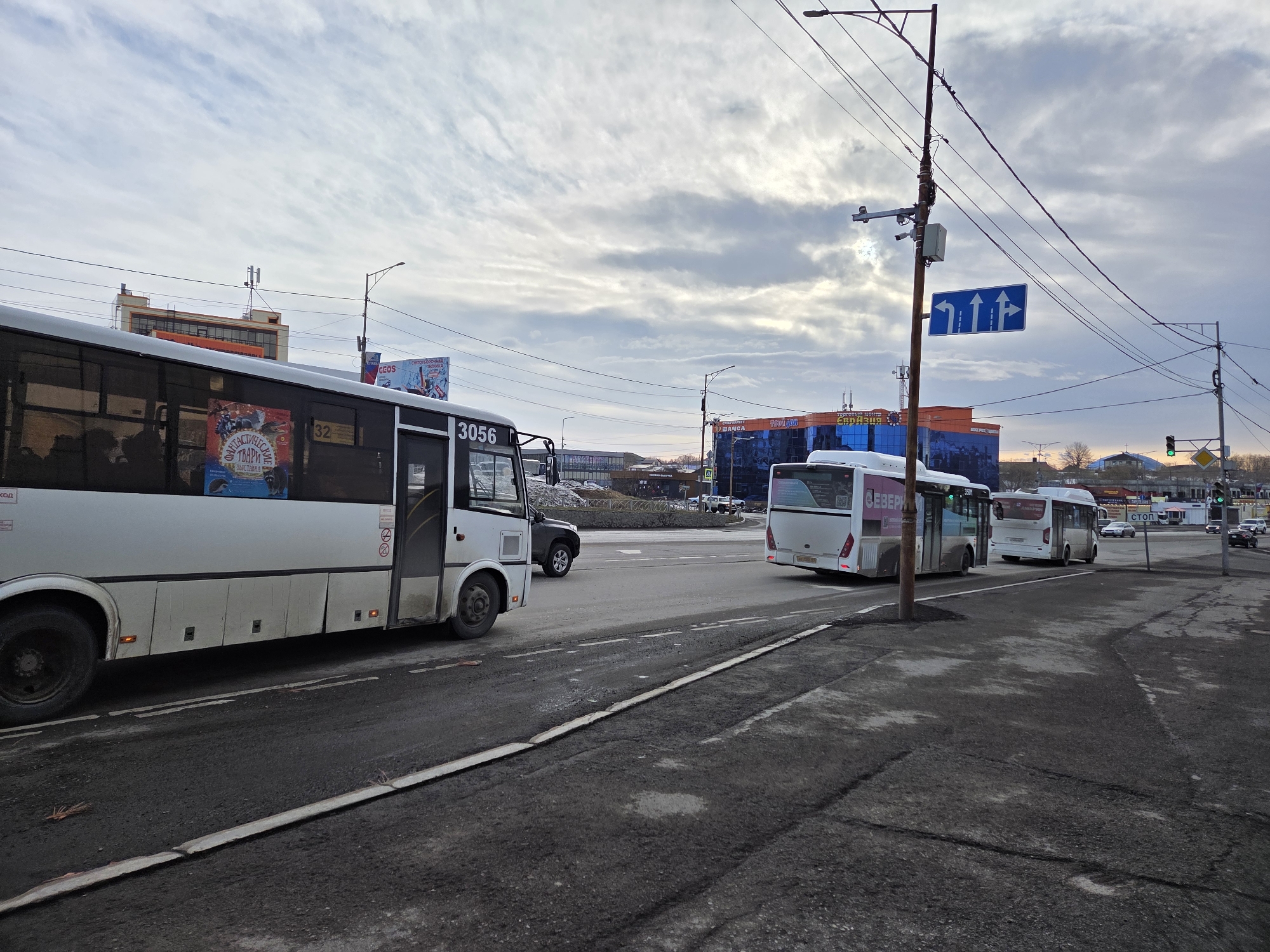 The image depicts a city street in an unknown location, featuring several buses. A white bus, numbered 3056, is positioned on the left side of the road. A black SUV follows behind. To the right of this bus, two more white buses are visible. A traffic sign indicating lane directions is mounted on a pole. The road is paved with asphalt, showing signs of wear and patching. Buildings are visible in the background. The sky is overcast, and streetlights are present. Other vehicles are seen in the distance.