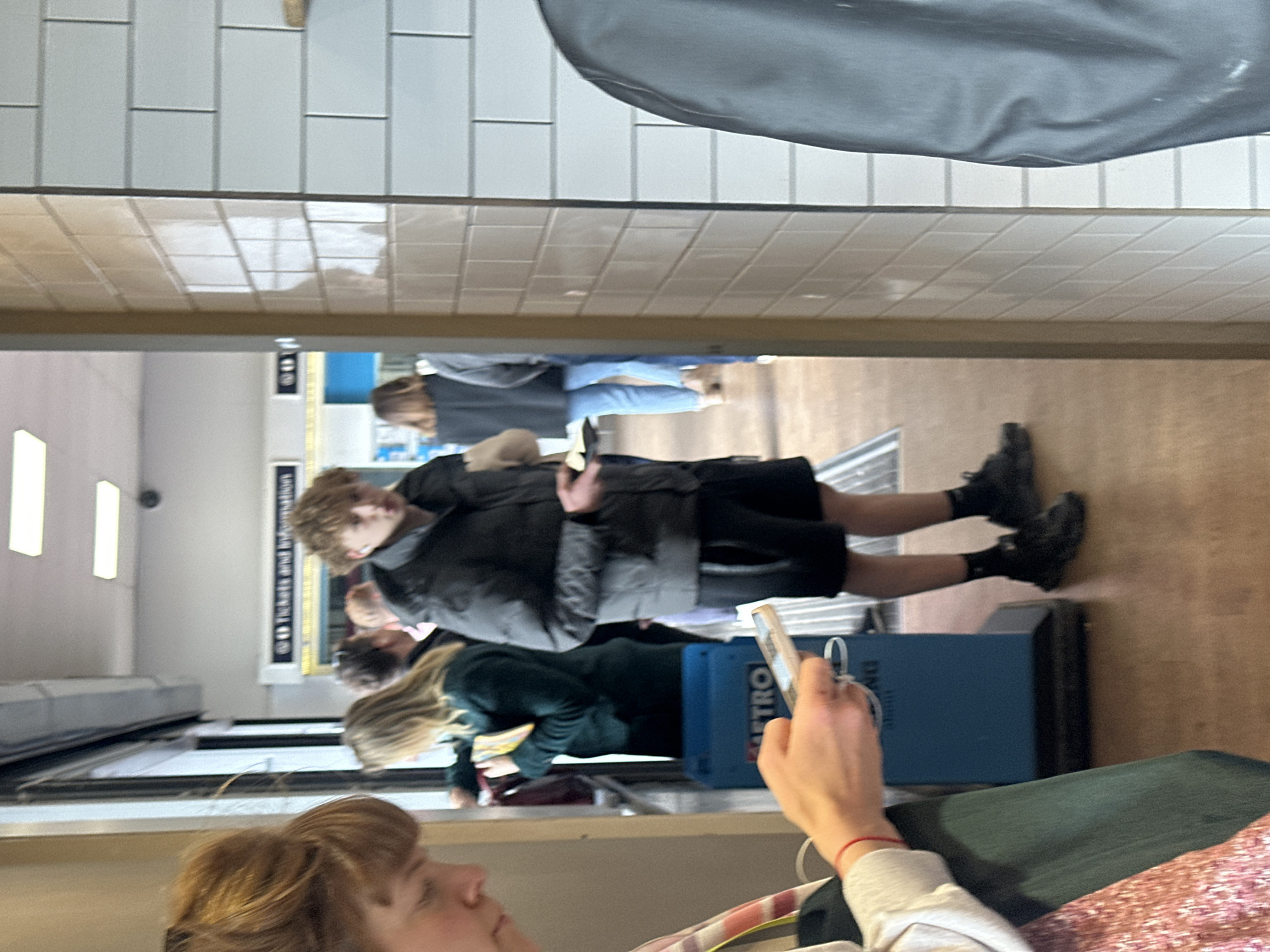 The image appears to be taken indoors, likely in a subway station in Tonbridge and Malling, United Kingdom. Several people are visible, primarily standing in line. One person is holding a phone, possibly taking the picture. Another individual is wearing a dark grey jacket, black shorts, and black shoes. A blue box with "METRO" printed on it is also in view. The background shows a white tiled wall and a sign that may say "Tickets and Information." The overall environment seems like a typical waiting area at a subway station.