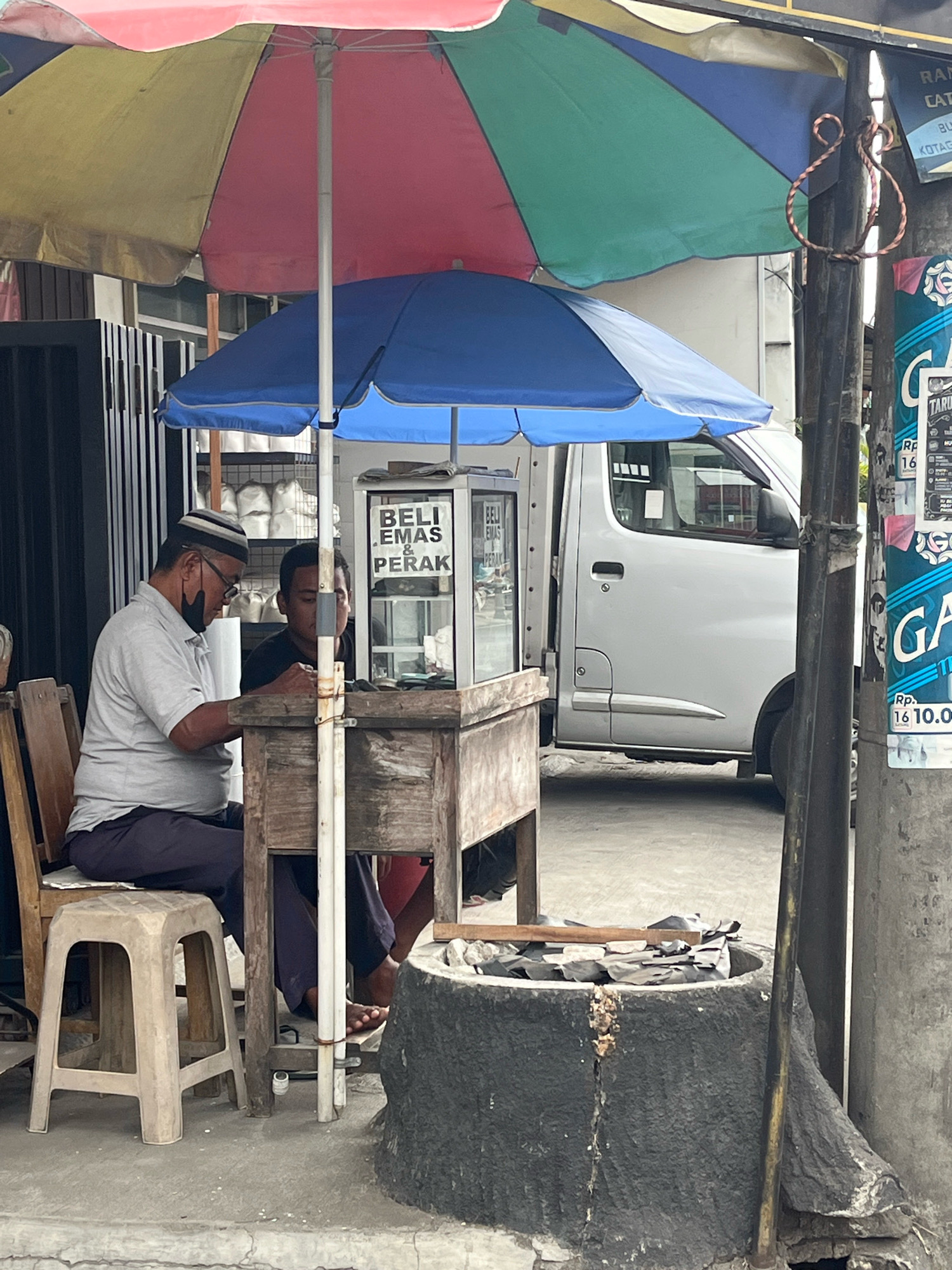 Two men are seated at a makeshift stall under colorful umbrellas. The man on the left, wearing a light-colored shirt, dark pants, a peci cap, and a black mask, is looking down at a table. He is sitting on a plastic stool. The man on the right, wearing a dark shirt, is partially visible behind the table, also looking down. The stall features a glass display case with text that reads "BELI EMAS & PERAK" (Buy Gold & Silver). In the background, a white van is parked. A concrete structure, possibly a well or planter, is in the foreground. A utility pole with posters is to the right of the scene. The overall environment suggests a street-side business operation in a city setting.