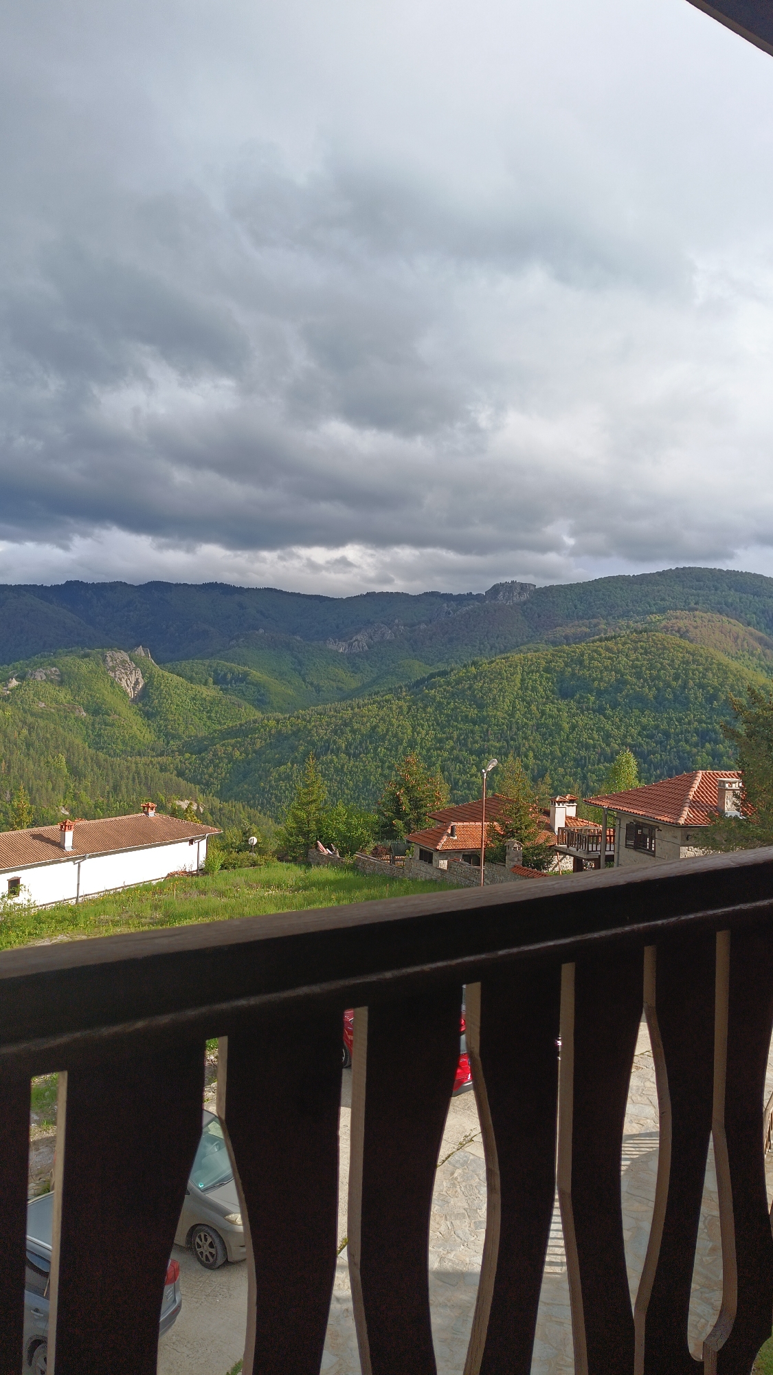 View from a balcony featuring a landscape of tree-covered mountains under a cloudy sky. In the midground, there are houses with terracotta tile roofs. A wooden railing is visible in the foreground, partially obscuring a car parked below. The location is Vratsa, Bulgaria.