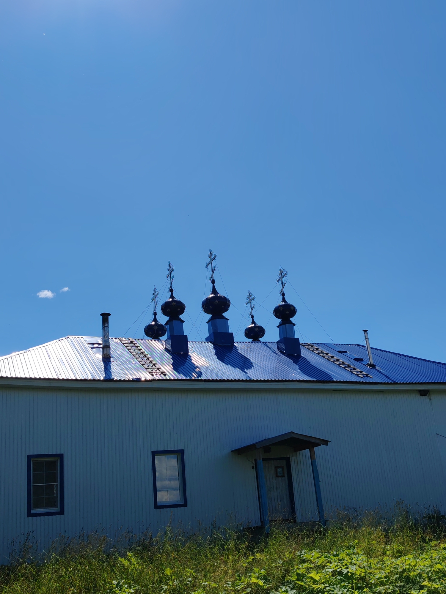 This is a vertical, medium-shot, outdoor photograph taken on a sunny day. The subject is the side and roof of a white, wooden building, likely a church, under a clear blue sky with a few wispy clouds. The building has vertical siding and two rectangular windows with dark blue trim. A small, covered entrance with a wooden door is visible on the right side. The roof is a bright blue, corrugated metal. Several dark blue onion domes topped with crosses rise from the roof, characteristic of Russian Orthodox architecture. A metal chimney pipe stands on the left side of the roof, and a ladder is visible extending up the roof slope near the center. The foreground is filled with lush green grass and foliage. The scene is located in the city of Chuchlem, Russia.