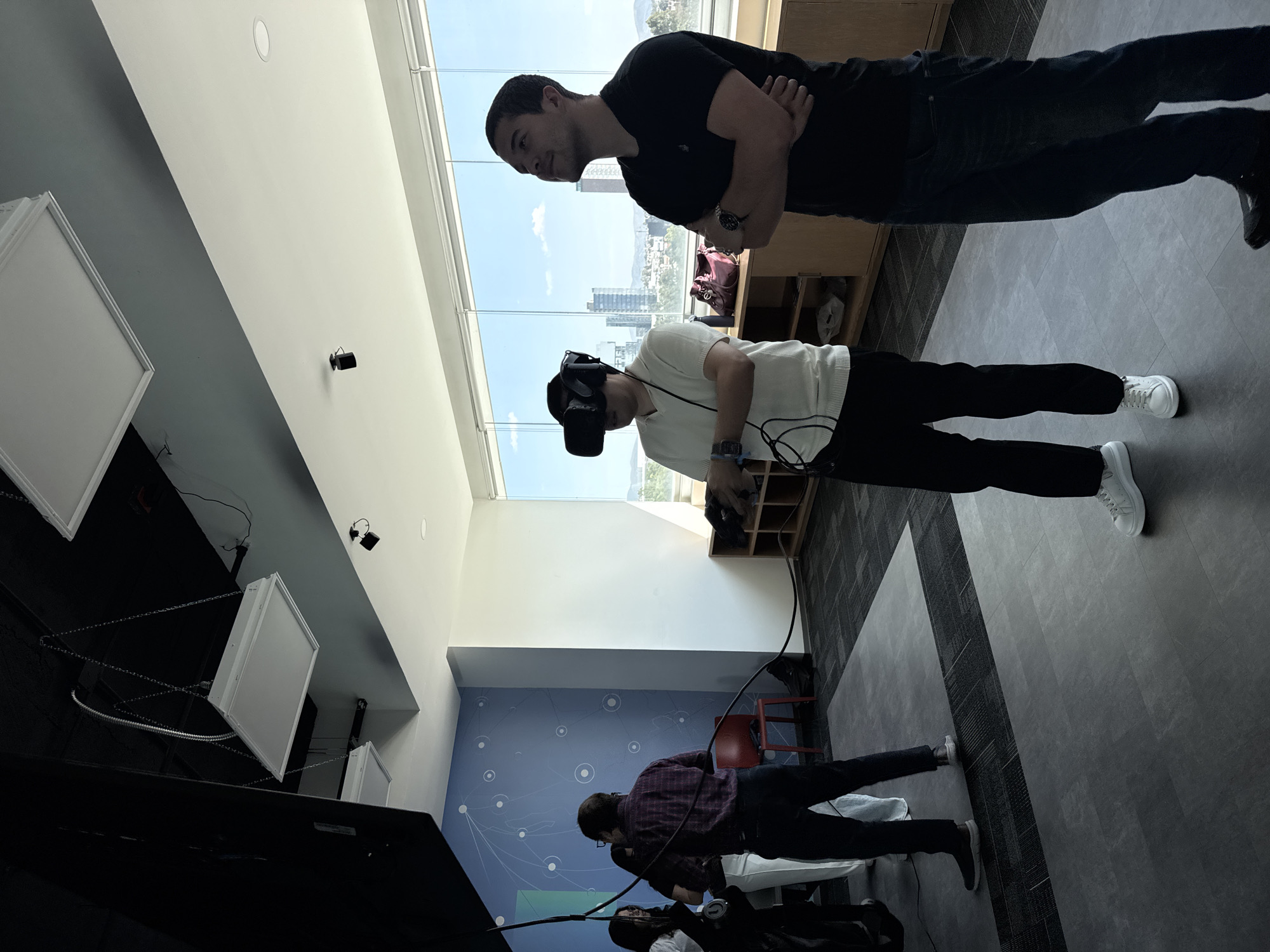 The image depicts an indoor scene with four people in what appears to be a modern office or event space. One person is wearing a VR headset and holding a controller, interacting with a virtual environment. Another individual stands nearby with crossed arms, observing. A third person, wearing a plaid shirt, seems to be either operating or watching an additional piece of technology. A fourth person is partially visible near the bottom of the image.
The location has large windows overlooking a cityscape with high-rise buildings, suggesting a location within an urban setting. Given the specific request to identify if the location is Puebla, Mexico, and based on an analysis of the visible buildings, the skyline, and the architectural style observable through the window, it cannot be definitively confirmed that this image depicts Puebla, Mexico. The architectural styles do not provide definitive evidence of Puebla, Mexico. Without additional visual cues or metadata, such as signage or geolocation tags, a conclusive identification is not possible.
The room is lit by overhead lighting, including recessed lights. The walls are painted in white and have a blue accent wall with a pattern of circles and lines. There is a wooden shelf unit visible, along with seating and equipment. The floor is a combination of carpeted and tile flooring.