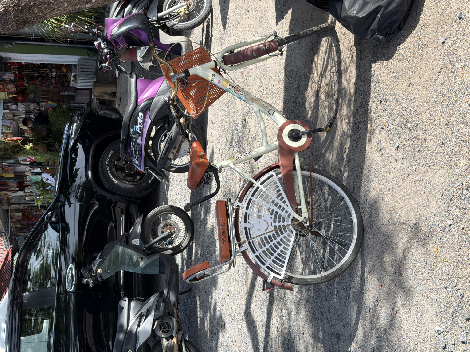 In Baan Plai Laem, Thailand, a white bicycle with brown seats and trim is parked on a gravel surface. The bicycle has an orange basket on the handlebars and a white, web-like design on the rear fender, which includes a red and white smiley face. A black SUV is parked to the left of the bicycle, and two scooters are visible behind it. In the background, there are market stalls with various items displayed. The scene is illuminated by sunlight, casting shadows on the ground.