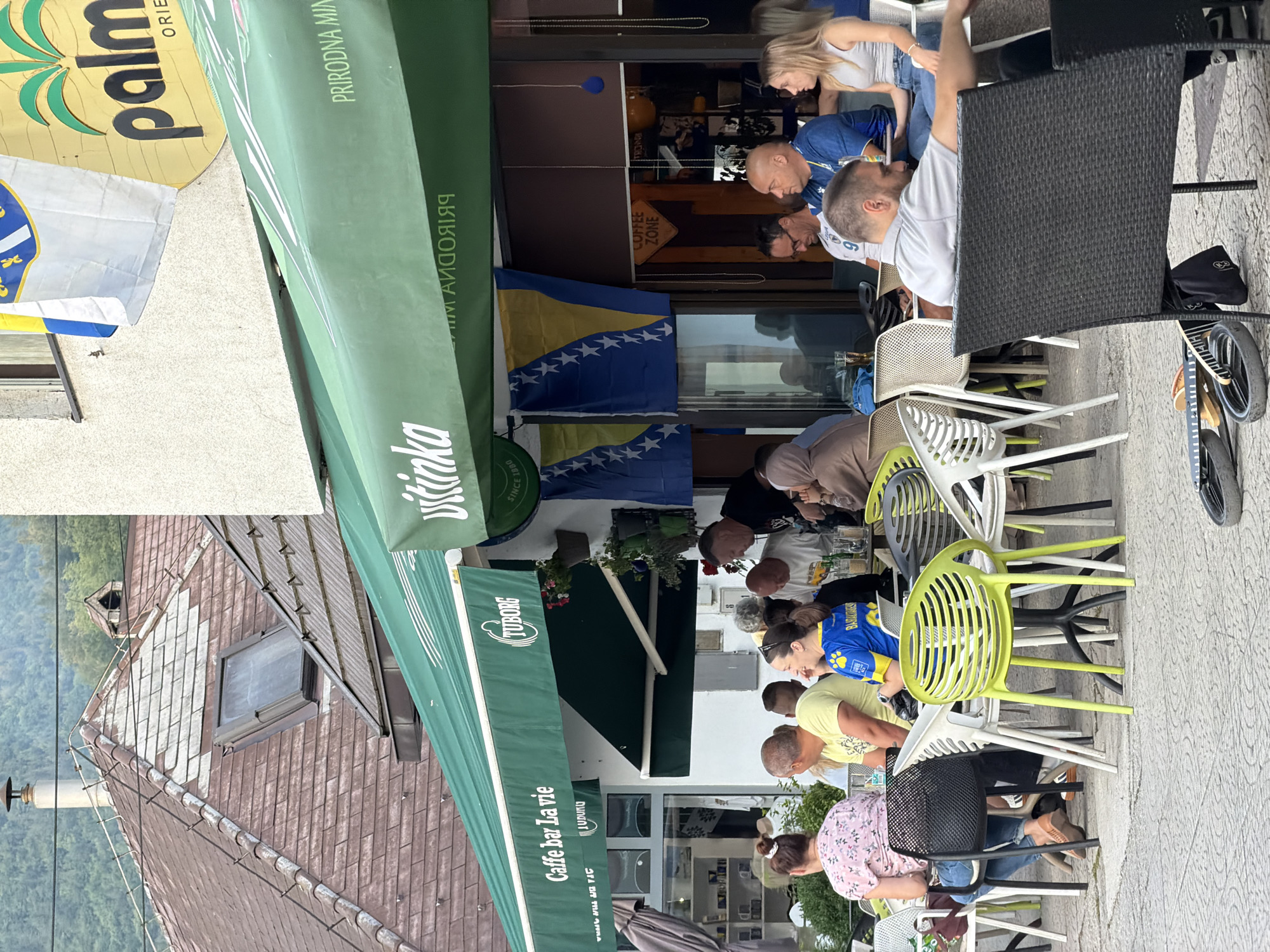 The image depicts an outdoor seating area of a cafe, identified as "Caffe bar La vie" with a Tuborg advertisement, in Travnik, Bosnia and Herzegovina. Several patrons are seated at tables. In the background, a building with a tiled roof and a skylight is visible, along with a green awning extending over the cafe's seating area. Two flags, one appearing to be the flag of Bosnia and Herzegovina, are displayed on the building. Various chairs of different colors and materials are arranged around the tables. Some people are engaged in conversation, while others are looking towards the camera or elsewhere. The ground is paved with patterned tiles.